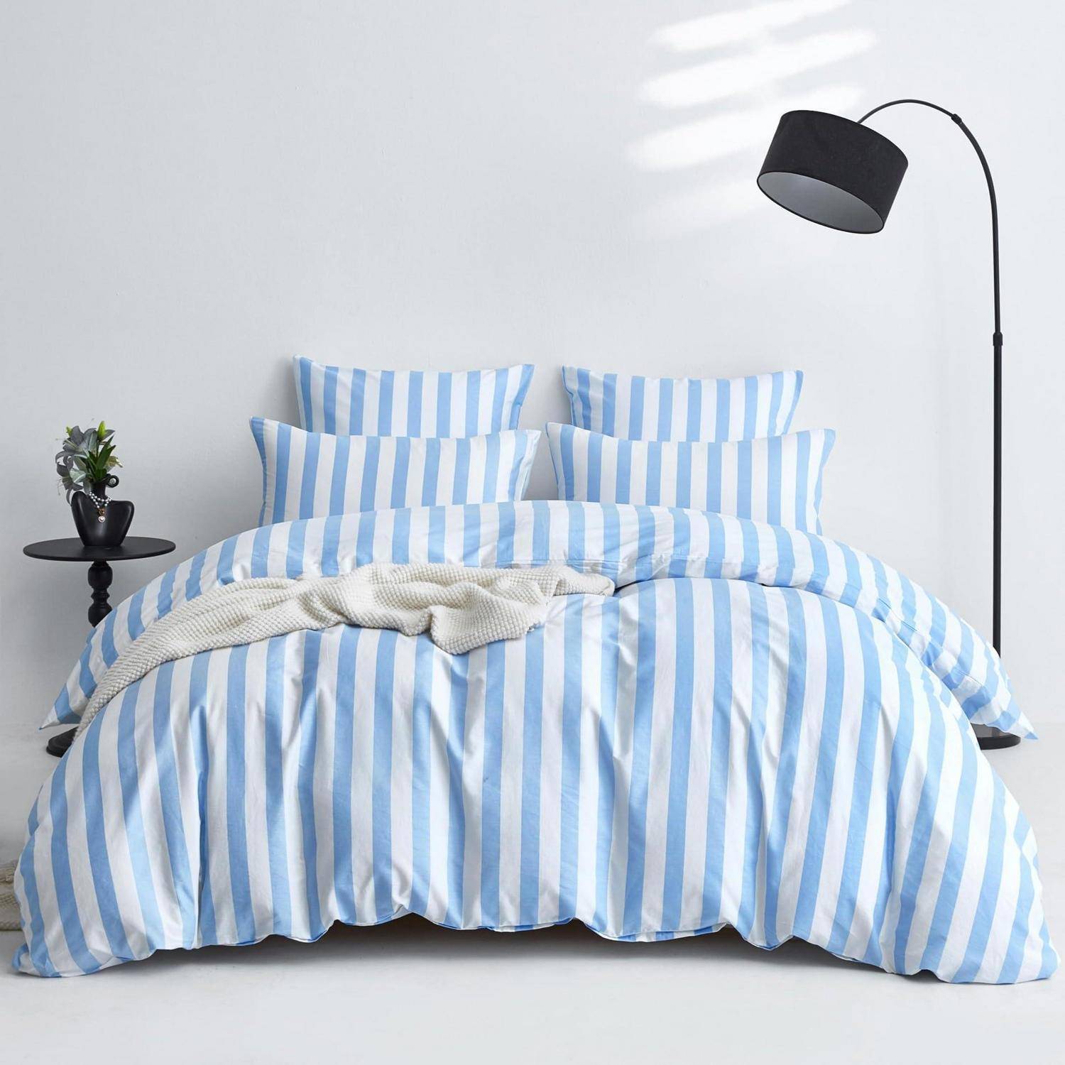 Gestreiftes blau-weißes Bettzug auf einem ordentlich gemachten Bett mit einer Wurfdecke, neben einem kleinen Nachttisch und einer schwarzen Stehlampe.