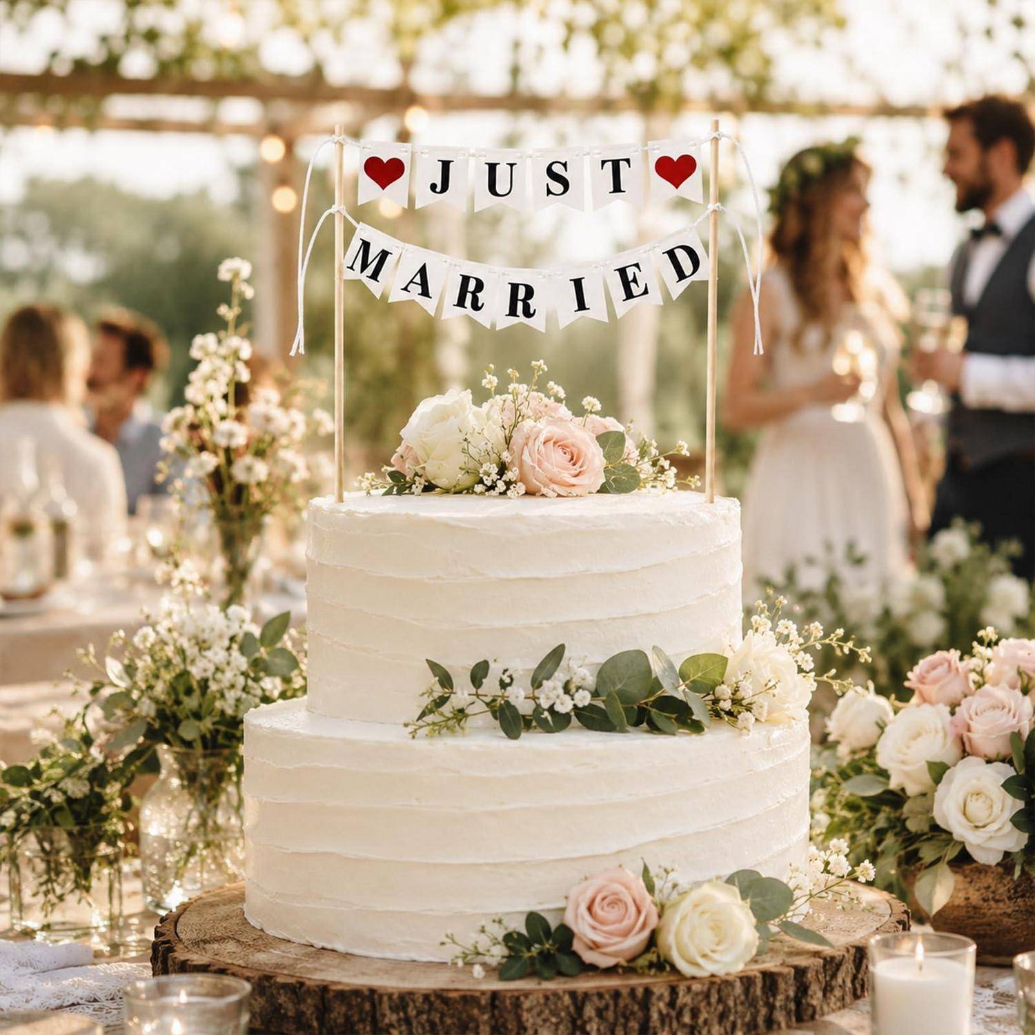 Weiße mehrstöckige Hochzeitstorte mit "JUST MARRIED""-Topper und Blumen, ausgestellt auf einem dekorierten Empfangstisch im Freien.