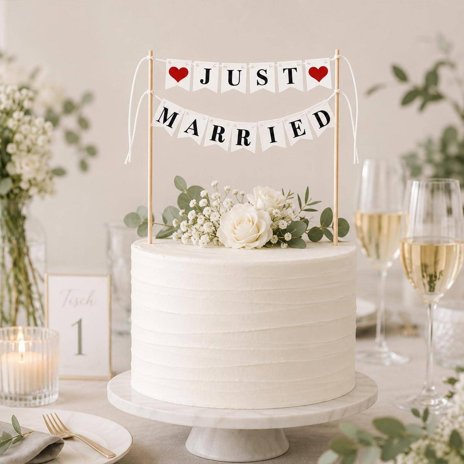 Weißer Hochzeitstorte auf einem Ständer mit 'JUST MARRIED' Wimpelkette und Blumentopfaufsatz, angerichtet auf einem dekorierten Empfangstisch mit Kerzen und Champagnergläsern.