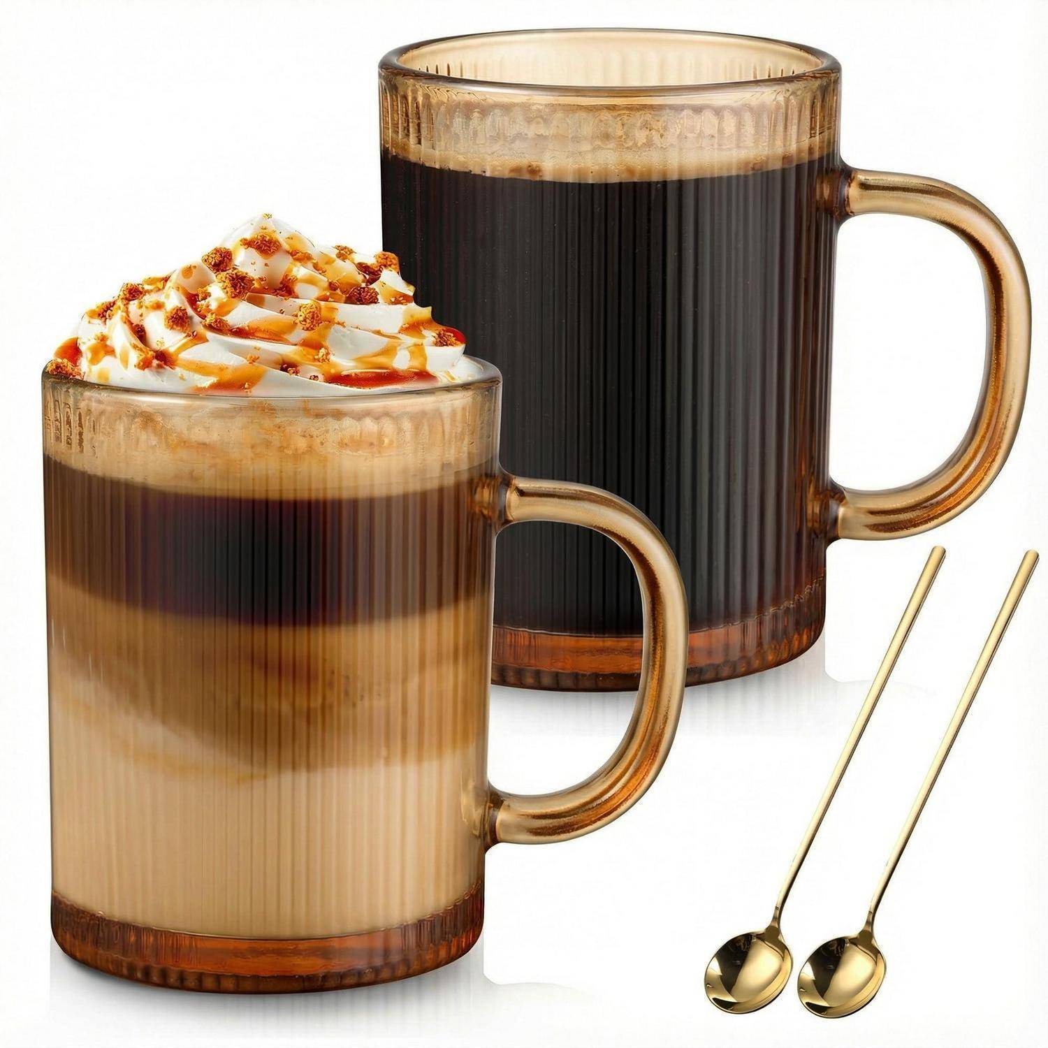 Zwei durchsichtige Tassen mit geschichteten Kaffegetränken auf weißem Hintergrund, eine mit Schlagsahne und Karamell-Drizzle garniert, mit zwei goldenen Löffeln daneben.