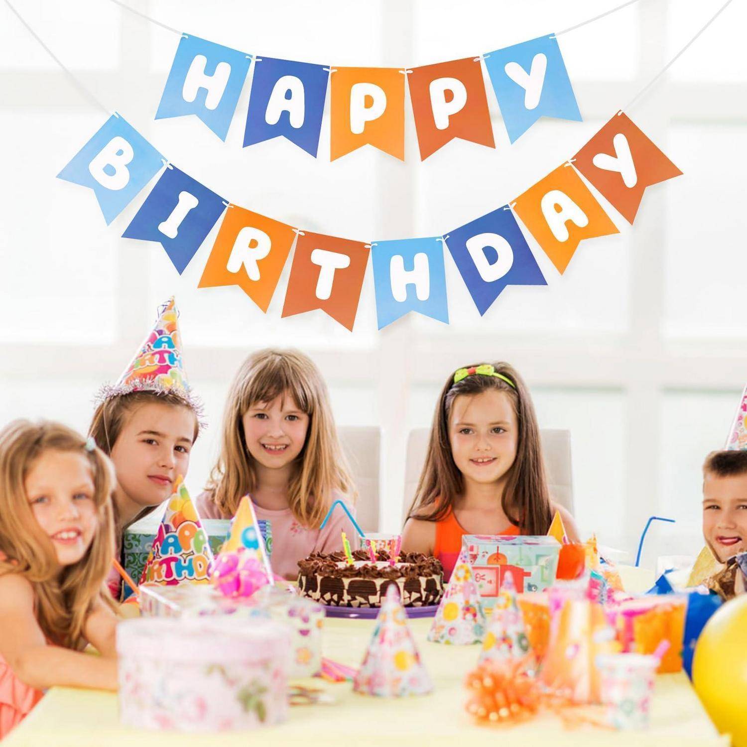 Kinder in Partyhüten sitzen um einen Geburtstagstisch mit Kuchen und Dekorationen unter einem „ALLES GUTE ZUM GEBURTSTAG"-Banner.