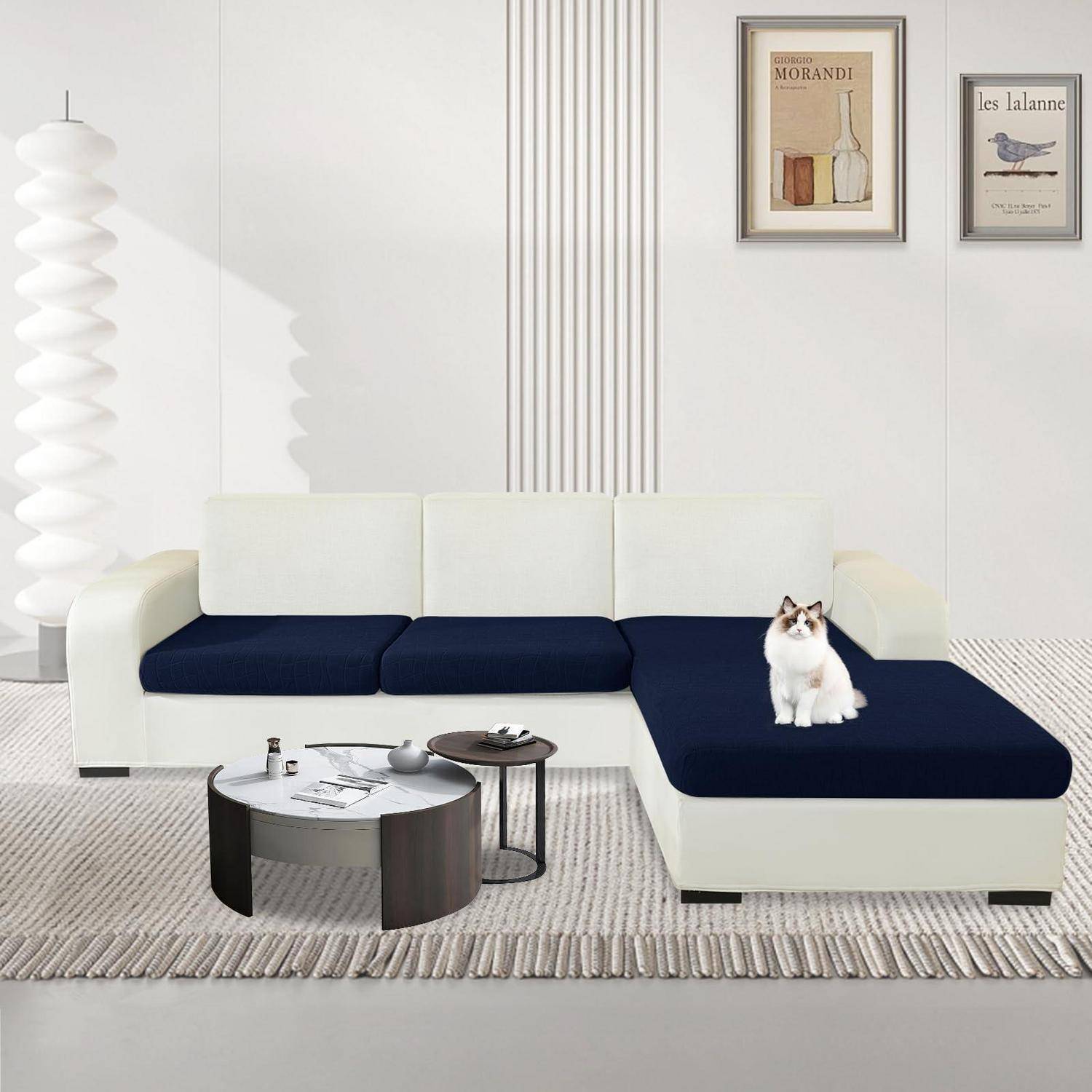 Modernes weißes Ecksofa mit dunkelblauen Kissen und einer Katze, die auf der Chaiselongue sitzt, in einem hellen, minimalistischen Wohnzimmer mit Wandbildern im Rahmen und Couchtischen.