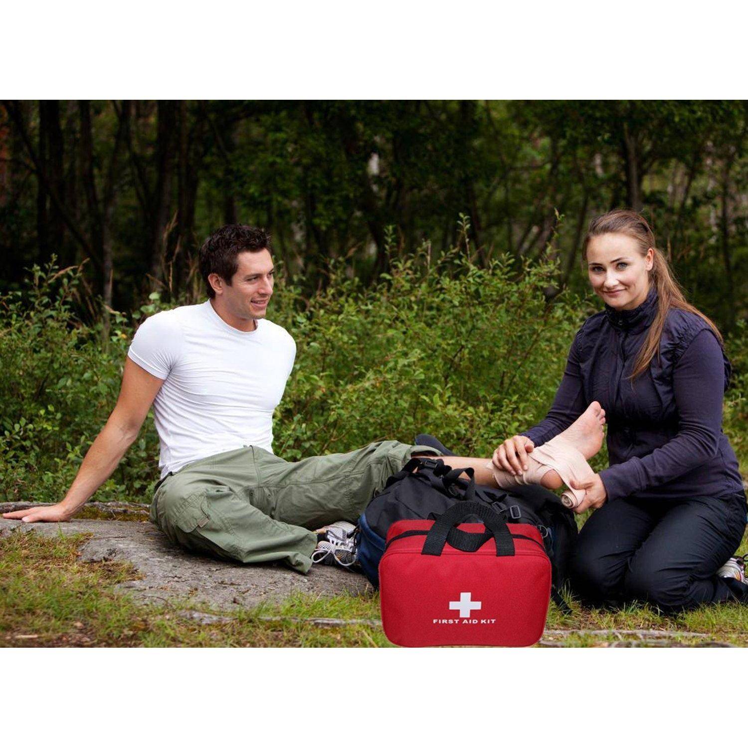 Zwei Personen führen draußen Erste Hilfe an einem verletzten Knöchel neben einer roten Tasche "ERSTE-HILFE-KASTEN"" durch.