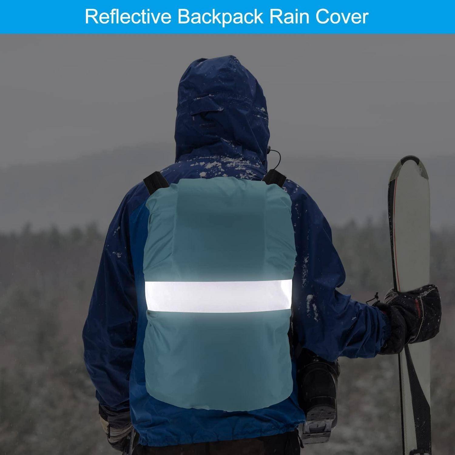 Person in blauer Winterkleidung, der eine reflektierende Regenschutzhülle für den Rucksack trägt, während er Skier hält, was die Sichtbarkeit unter Schneefall unterstreicht.