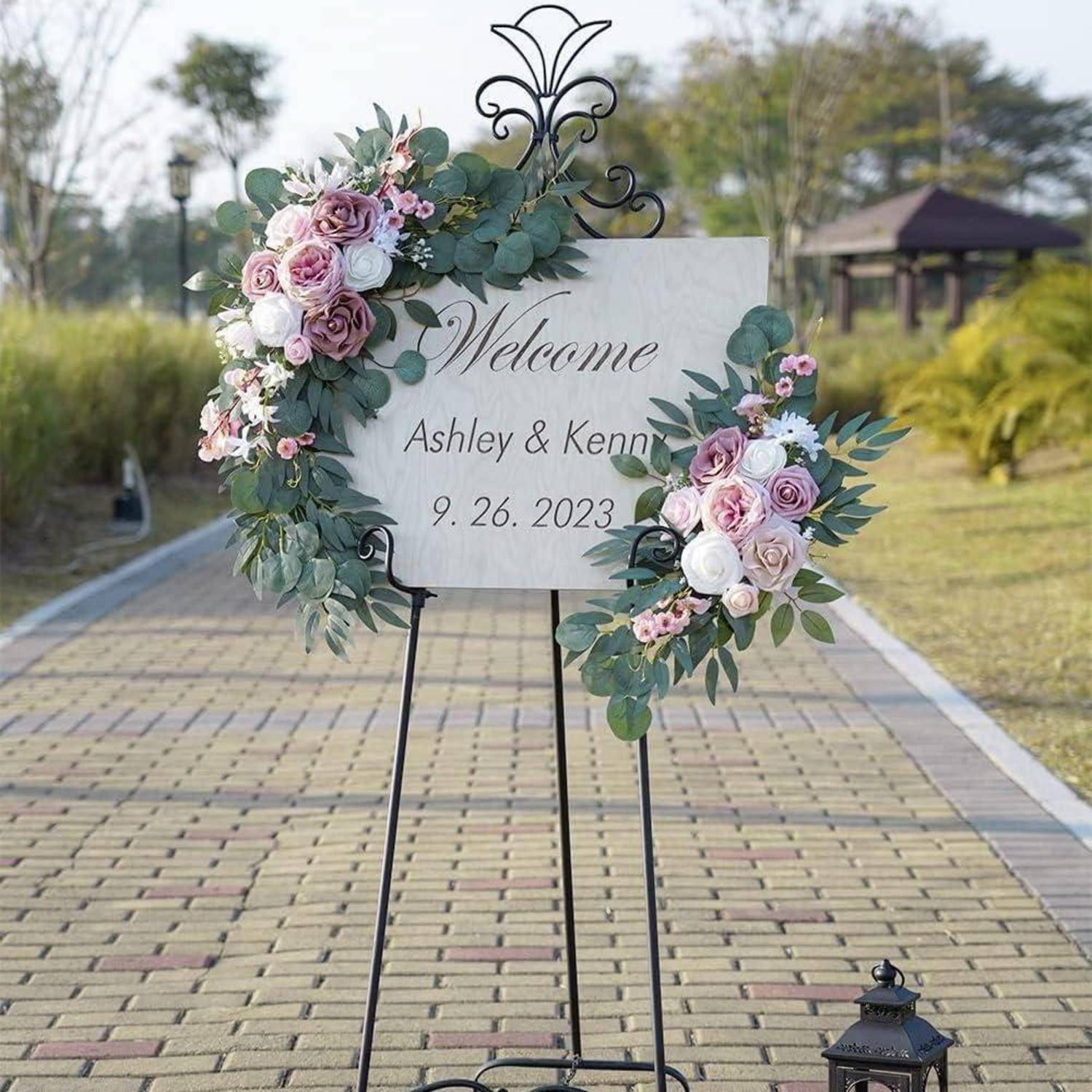 Willkommensschild zur Hochzeit mit Blumendekoration, auf dem „Welcome Ashley & Kenny 9.26.2023" steht, auf einem Ständer im Freien entlang eines Weges.