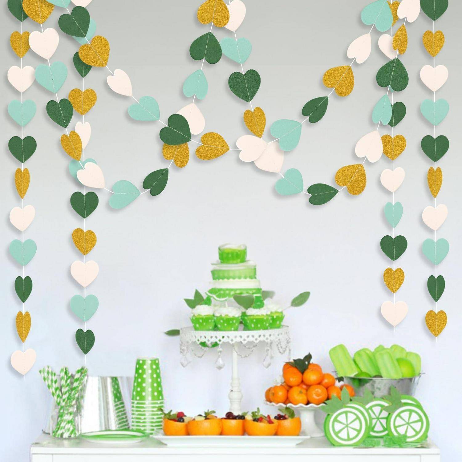Grüne mehrstöckige Torte auf einem weißen Ständer mit Obst und Getränken auf einem Tisch, darunter hängende Herzgirlanden in Grün, Gold und Weiß.