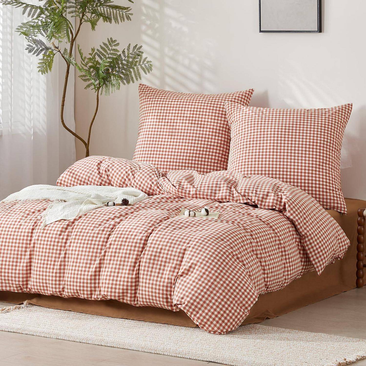 Bett mit rot-weiß karierter Bettwäsche und zwei passenden Kissen in einem hellen, minimalistischen Schlafzimmer mit einer Topfpflanze und Wandkunst im Rahmen.