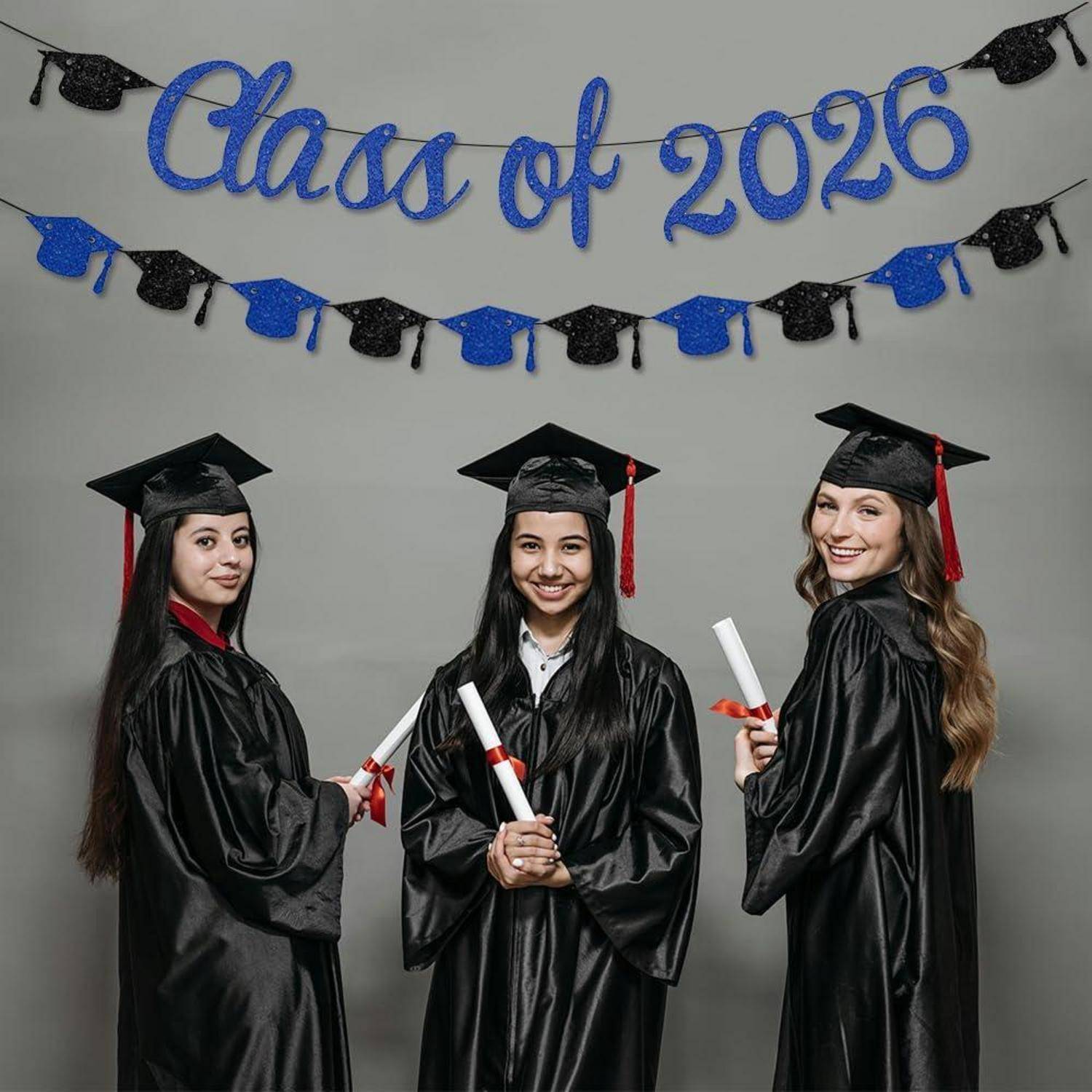 Drei Absolventen in Talaren und Doktorhüten posieren mit Diplomen in den Händen unter einem Banner mit der Aufschrift „Klasse von 2026