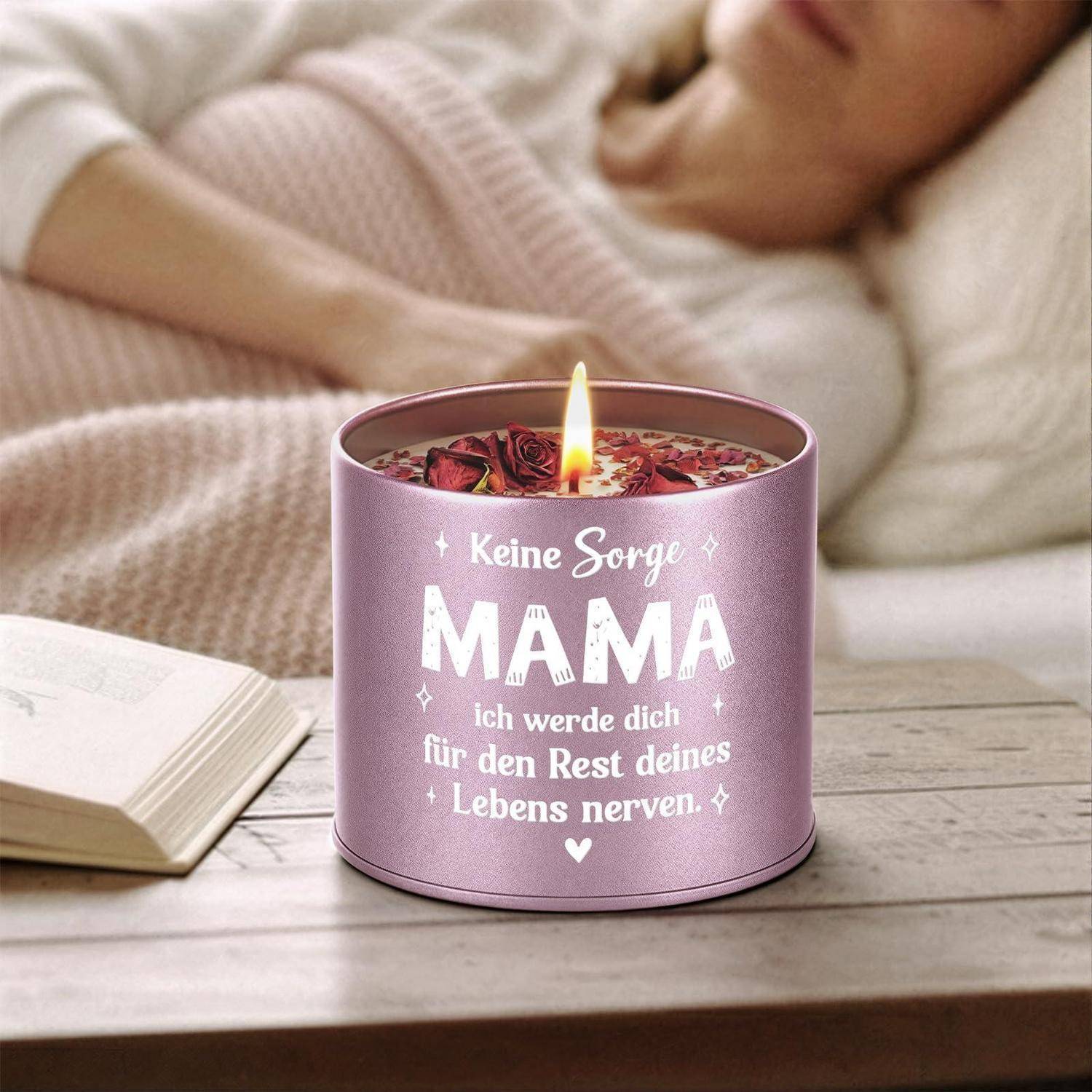 Rosa angezündete Kerze mit deutscher Nachricht „Keine Sorge MAMA, ich werde dich für den Rest deines Lebens nerven" auf einem Tisch neben einem offenen Buch, mit einer schlafenden Person im Hintergrund.