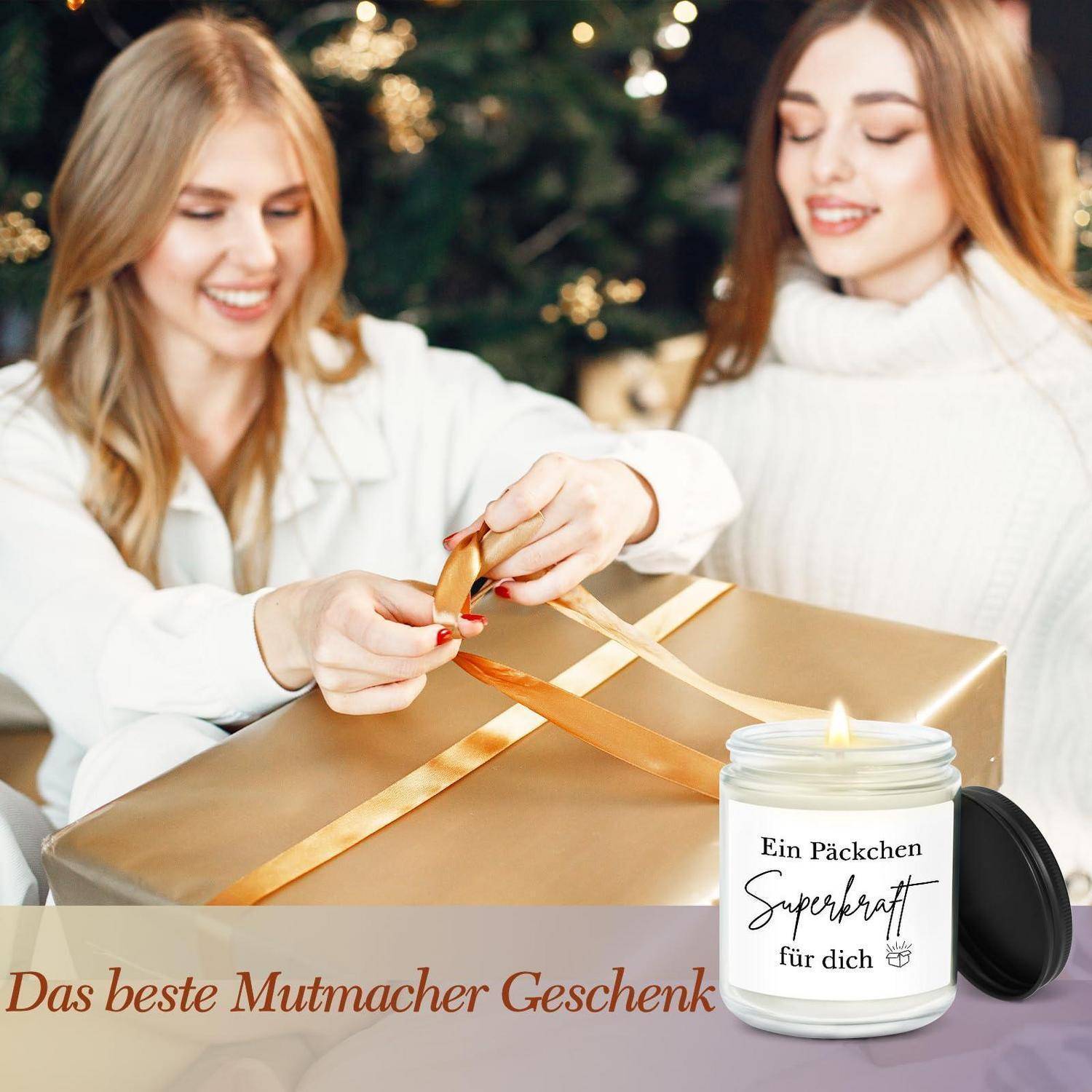 Zwei lächelnde Frauen in weißen Pullovern wickeln eine goldene Geschenkbox mit einem Band ein; ein Kerzenglaszug mit dem Etikett „Ein Päckchen Superkraft für dich" wird mit der Bildunterschrift „Das beste Mutmacher Geschenk"" gezeigt.