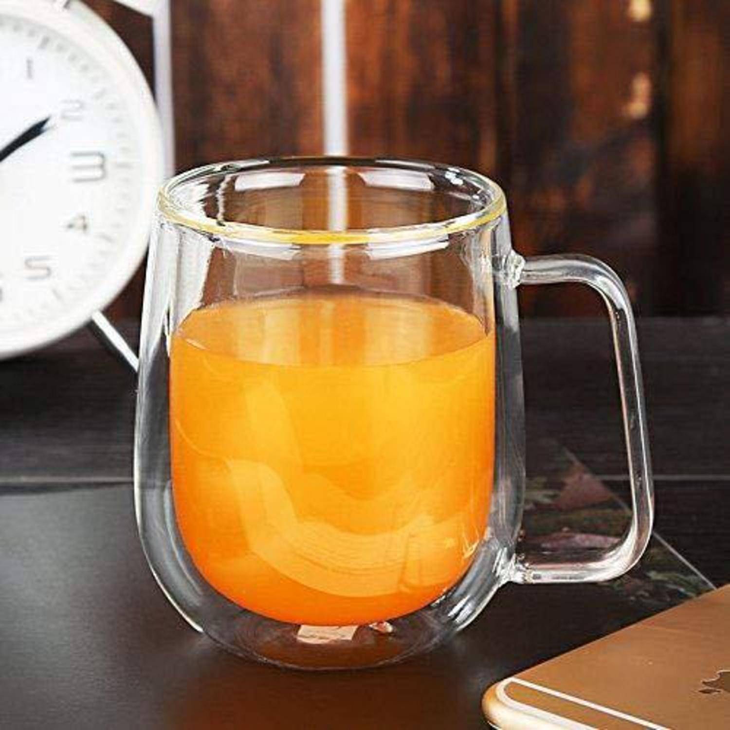 Ein klares doppelwandiges Glaseaus mit Orangensaft gefüllt steht auf einem Tisch, mit einer unscharfen Uhr im Hintergrund.