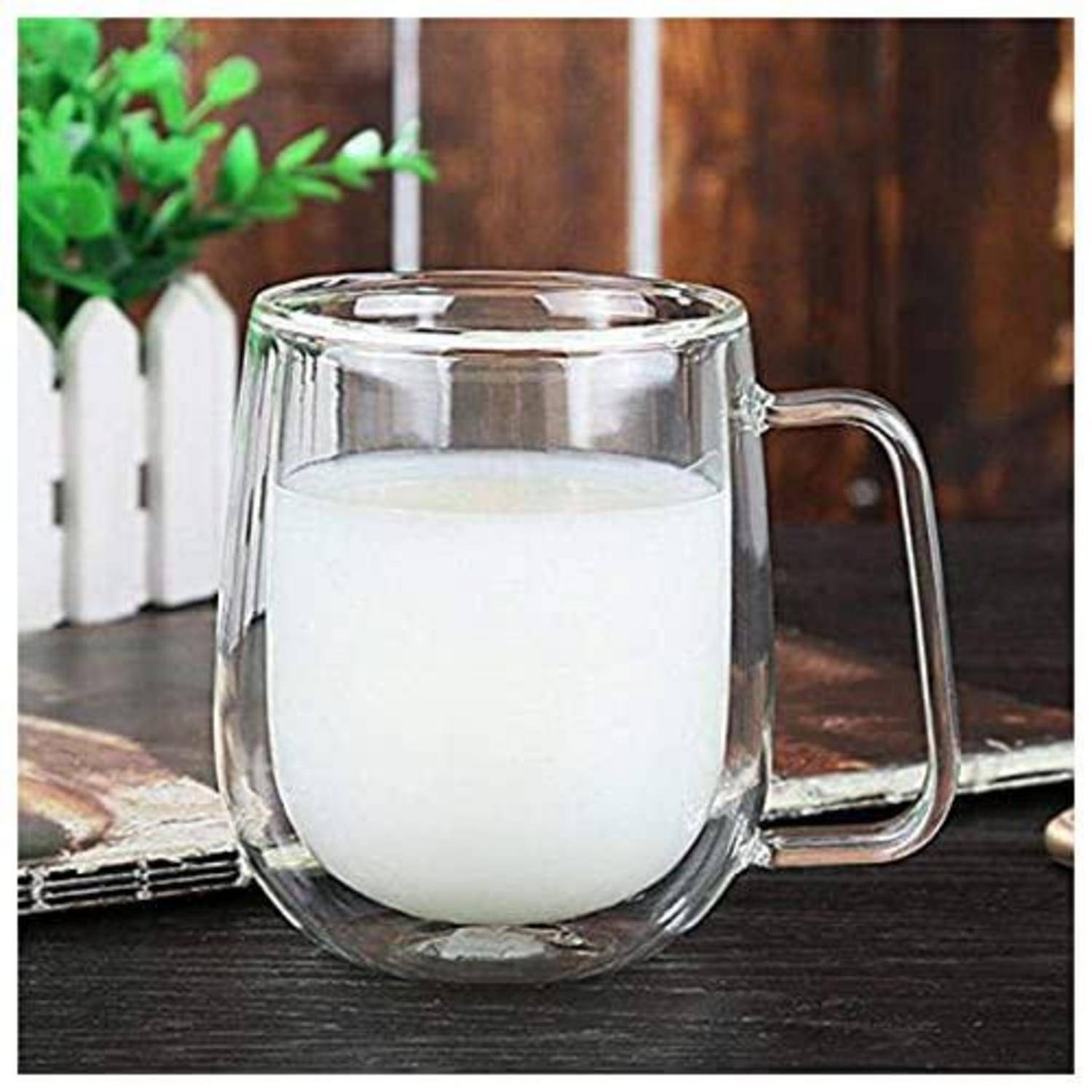 Ein klares doppelwandiges Glaskaffeetasse gefüllt mit Milch, gezeigt auf einem Tisch mit einer unscharfen Pflanze und Holzhintergrund.