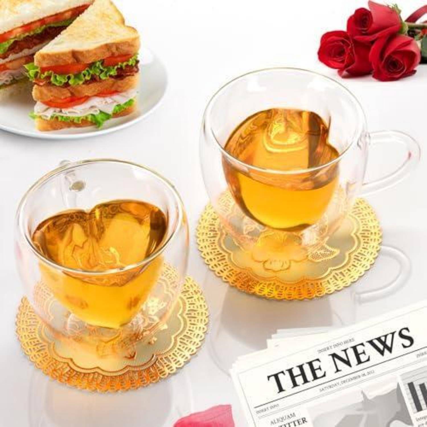 Zwei herzförmige Glastassen mit bernsteinfarbenem Tee auf goldenen Spitzentischdeckchen, mit einem Sandwichteller, roten Rosen und einer Zeitung auf einem weißen Tisch.
