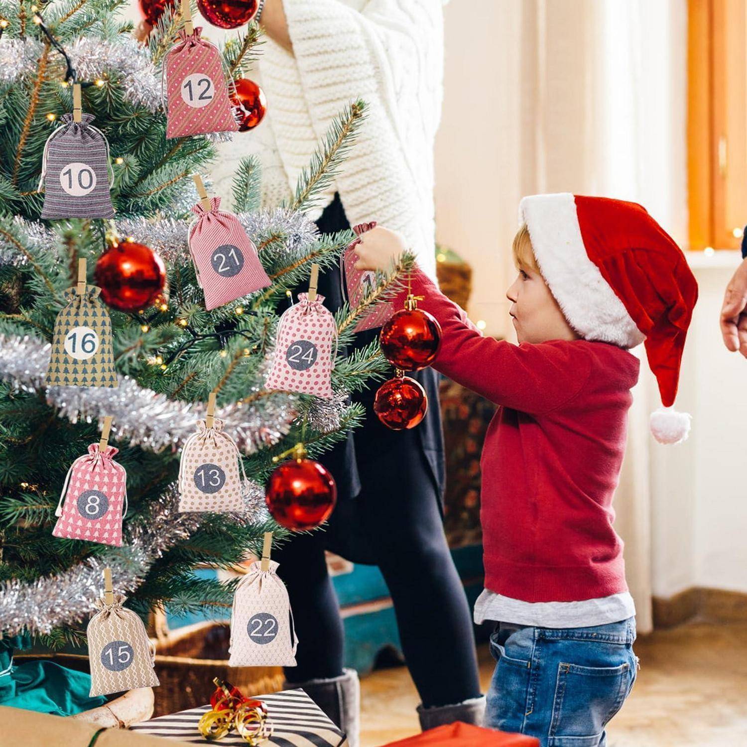 Junges Kind in einer Weihnachtsmütze hängt Ornamente an einen dekorierten Weihnachtsbaum mit nummerierten Geschenkanhängern auf, was auf eine Adventskalender-Countdown-Aktivität hindeutet.