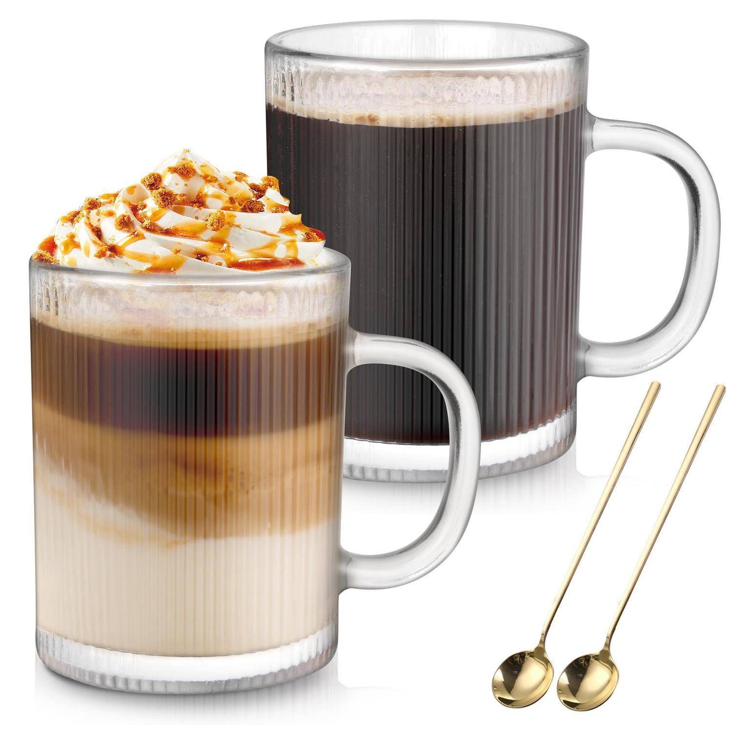 Zwei schichtige Kaffeegetränke in Glastassen, eines mit Schlagsahne und Karamellsirup garniert, mit zwei goldenen Rührlöffeln.
