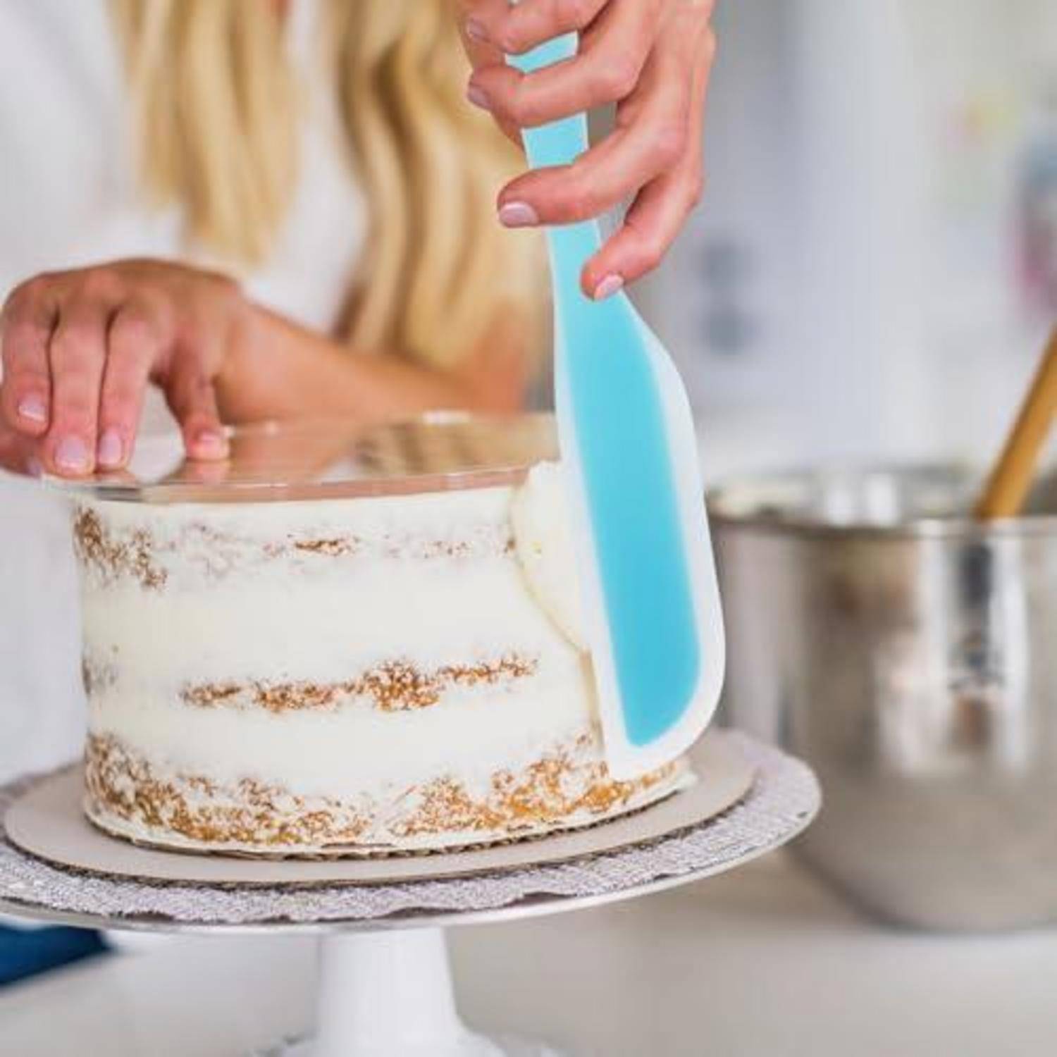 Eine Person glasiert einen mehrschichtigen Kuchen auf einer Drehplatte mit einem blauen Spachtel in einer Küche.