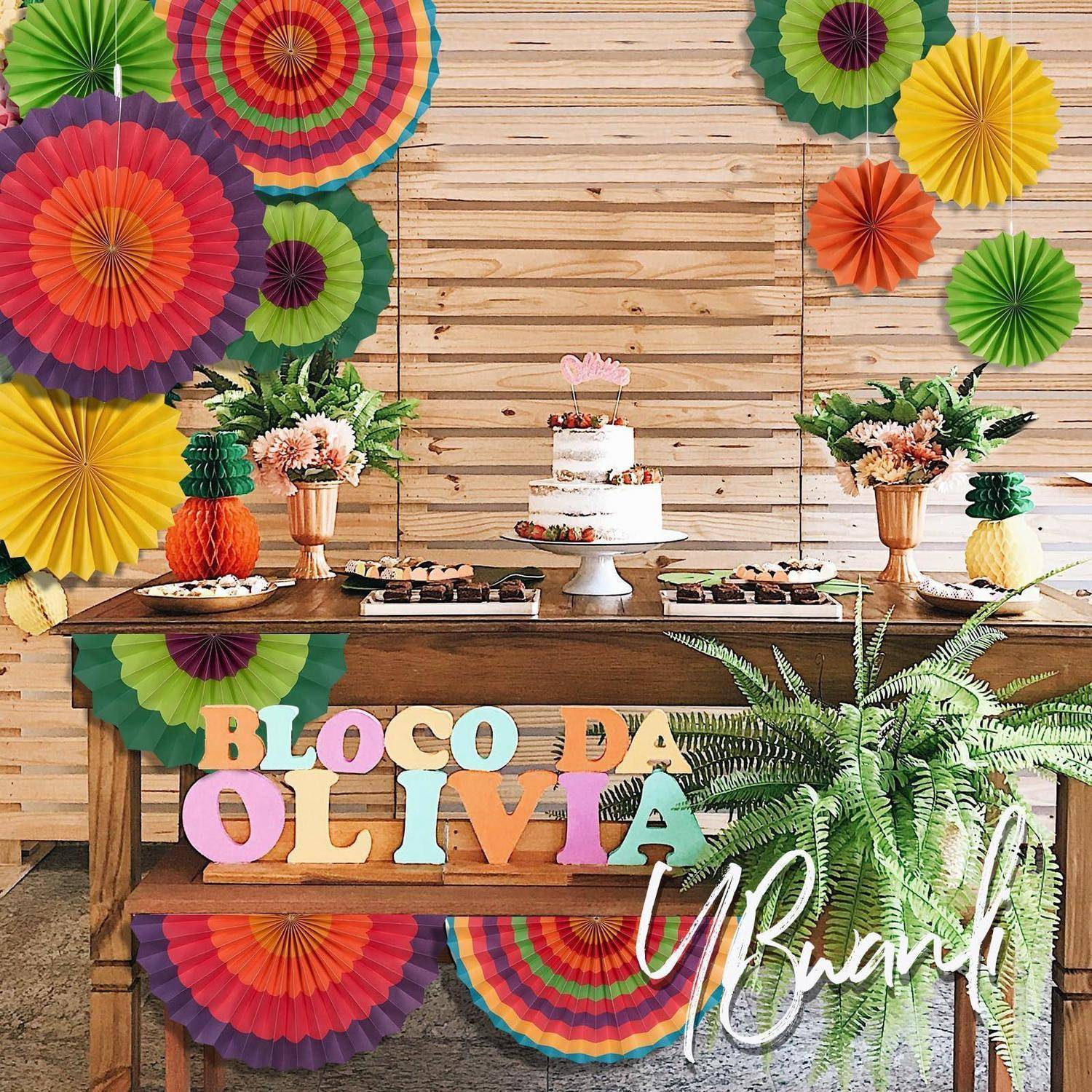 Farbenfroher Partydessert-Tisch für „BLOCO DA OLIVIA" mit Kuchen, Blumen, tropischen Dekorationen und bunten Papierfächern auf Holzhintergrund.