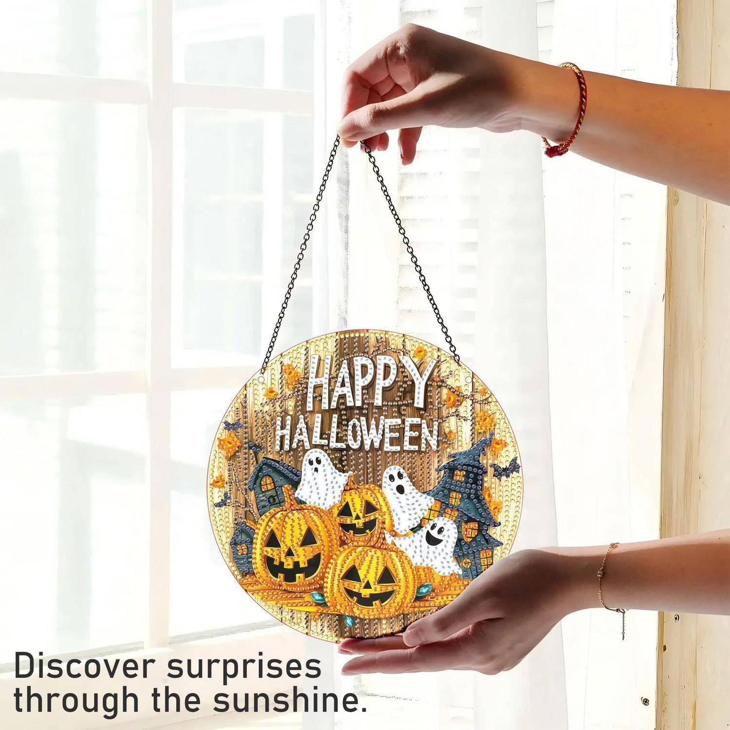 Hände halten ein rundes „HAPPY HALLOWEEN"-Hängschild mit Kürbissen, Geistern und einem Spukhaus gegen ein helles Fenster; der Text lautet „Entdecken Sie Überraschungen durch den Sonnenschein."