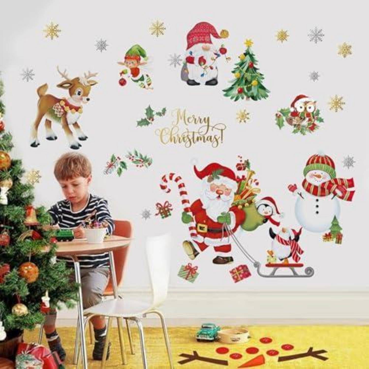 Ein kleiner Junge sitzt an einem kleinen Tisch neben einem geschmückten Weihnachtsbaum und liest oder schreibt, mit festlichen "Frohe Weihnachten""-Wandaufklebern, dem Weihnachtsmann, einem Schneemann, einem Rentier und Schneeflocken um ihn herum.