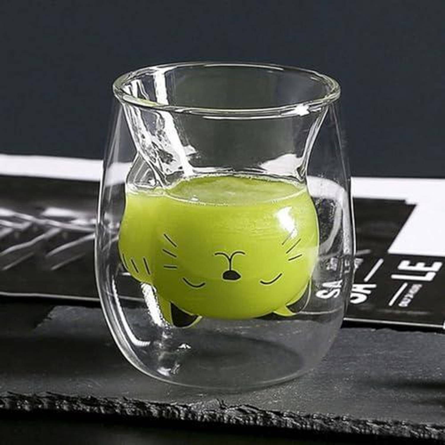 Eine grüne Cartoon-Gesichts-Tasse sitzt in einem durchsichtigen doppelwandigen Glasbecher auf einer dunklen Oberfläche.