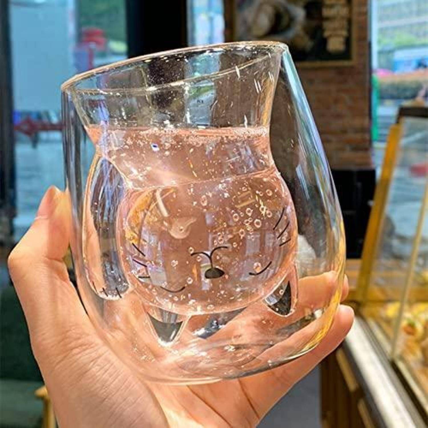 Eine Hand hält einen klaren, gebogenen Glasbecher mit einem gedruckten Cartoon-Katzenggesicht-Design vor einem Schaufenster.