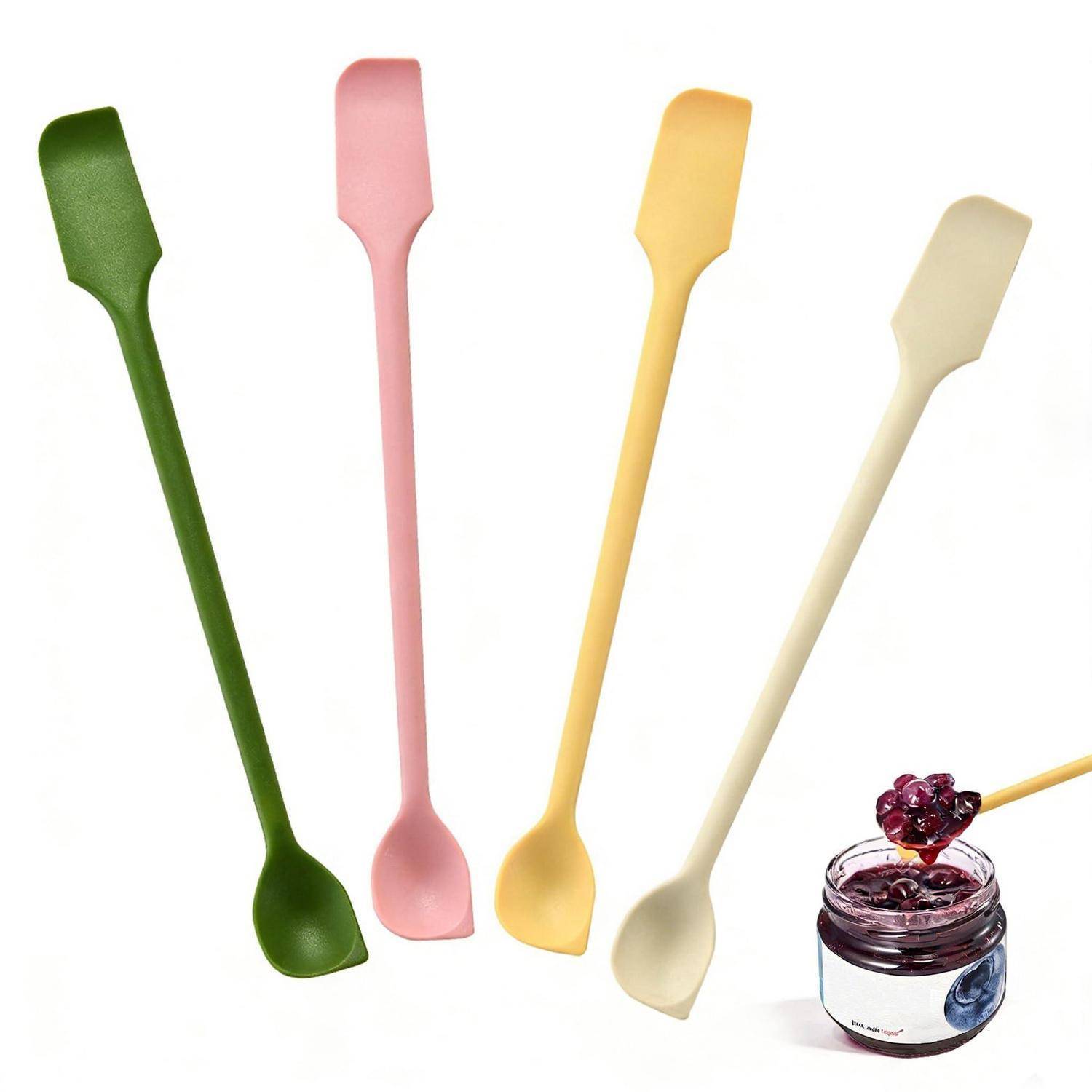 Vier lange Silikonmarmeladespachteln in Grün, Rosa, Gelb und Creme neben einem kleinen Marmeladenglas mit einem Löffel.