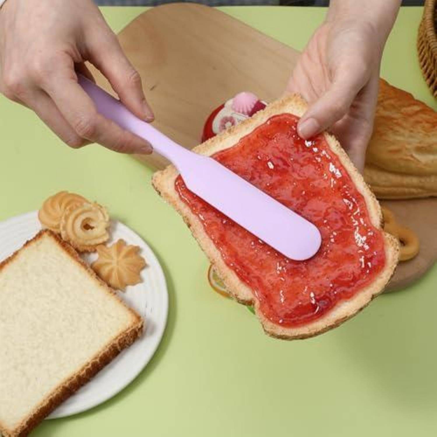 Hände verteilen Erdbeermarmelade auf eine Scheibe Weißbrot mit einem violetten Spachtel über einer hellgrünen Tischplatte, neben zusätzlichen Brotscheiben und Gebäck.