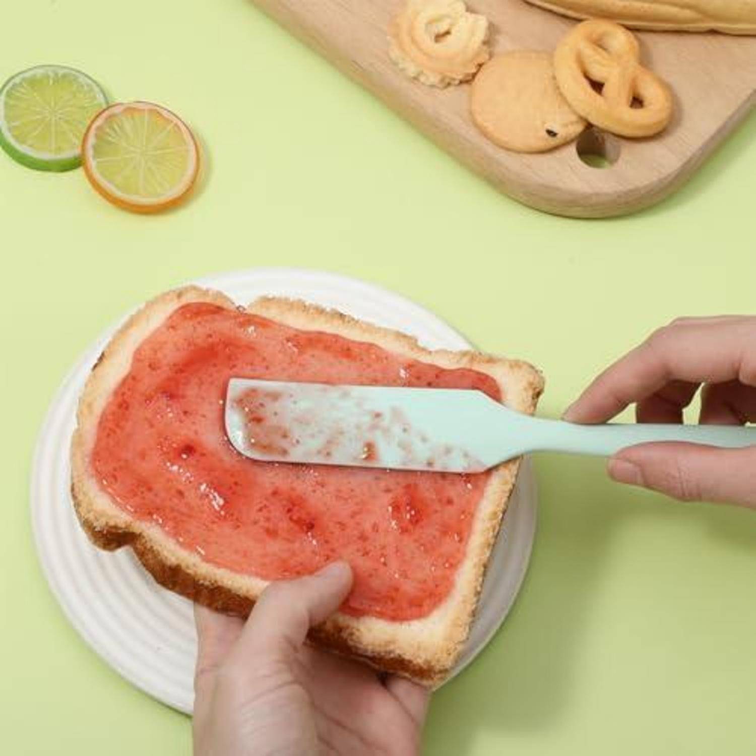 Hände verteilen rote Marmelade oder Soße auf einer Toastscheibe mit einem mintgrünen Spachtel; Limettenscheiben und Snacks liegen auf einem hellgrünen Tisch.