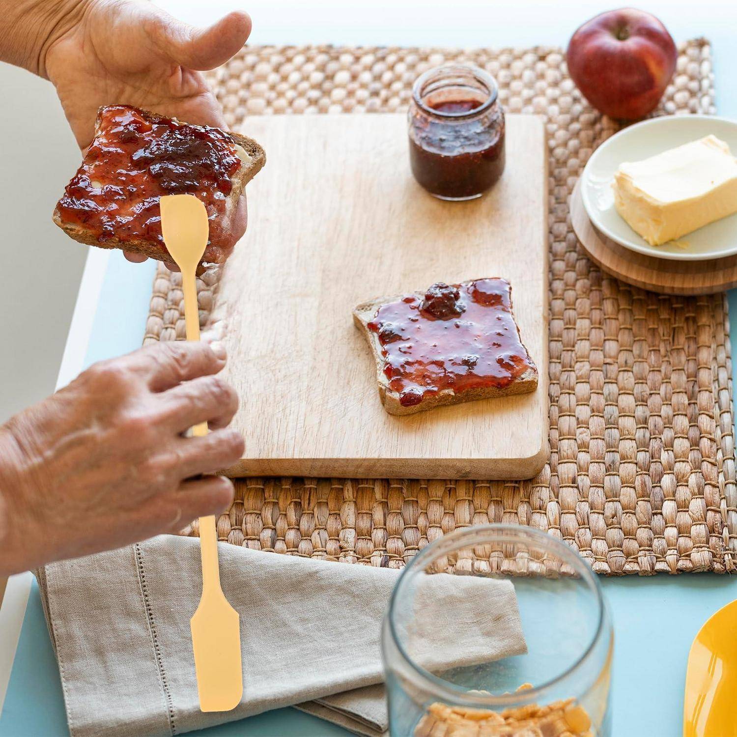 Hände verteilen Marmelade auf Toast über einem Holzschneidebrett, mit einem Glas Marmelade, Butter und einem Apfel auf einer Tischeinstellung.