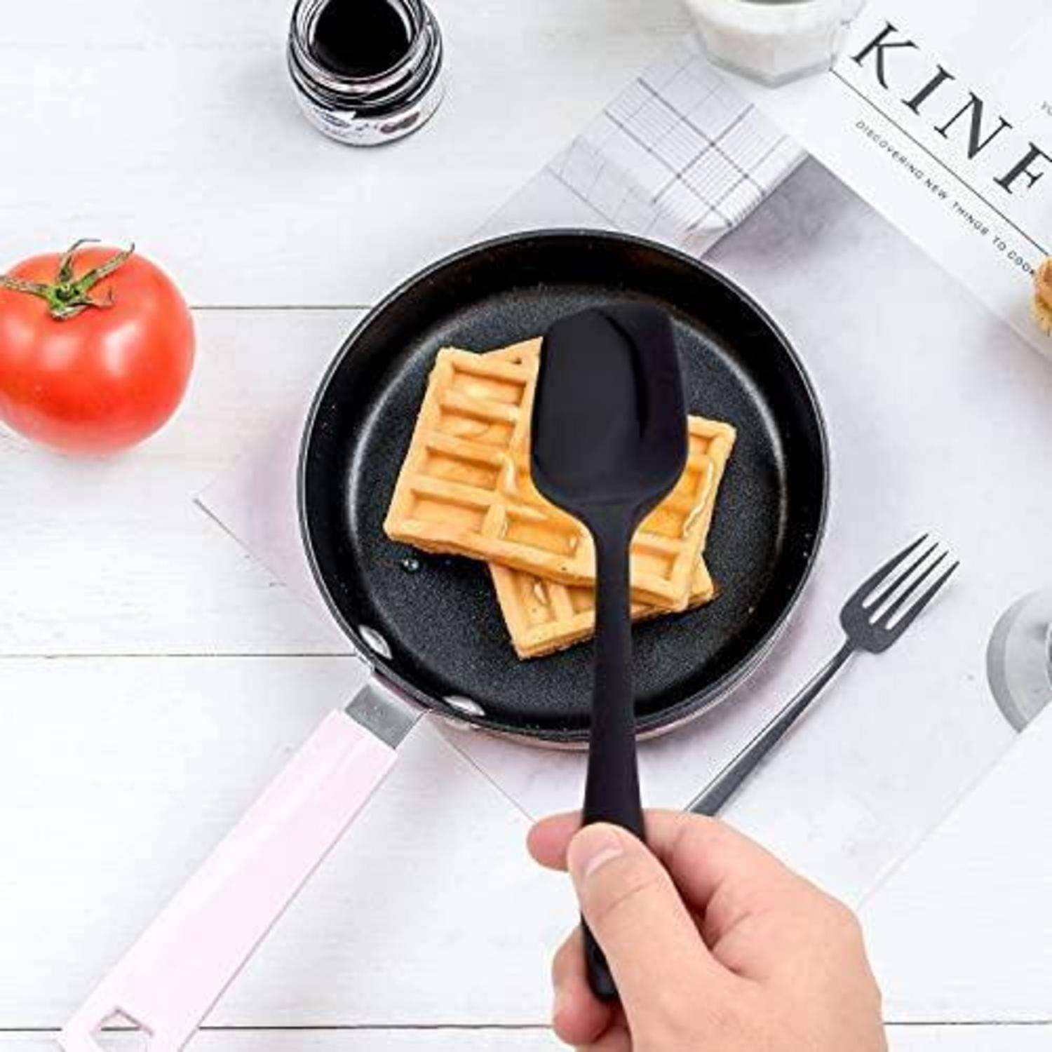 Eine Hand hält einen schwarzen Spatel über eine kleine Bratpfanne mit Waffelstücken auf einem weißen Tisch neben einer Tomate und einer Gabel.
