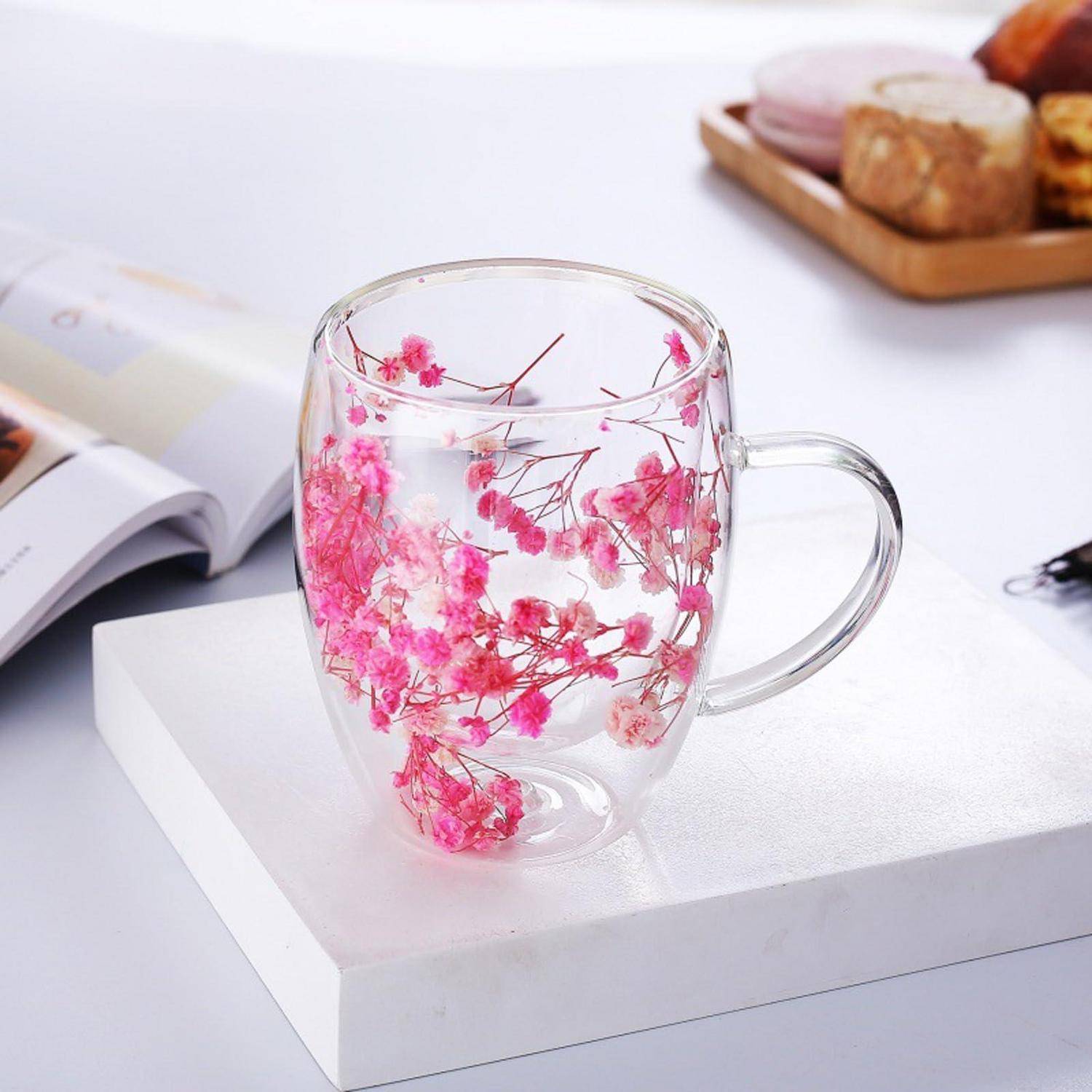 Klare Glastasse mit rosa getrockneten Blüten eingebettet im Inneren, ausgestellt auf einem weißen Sockel auf einer Tischplatte.