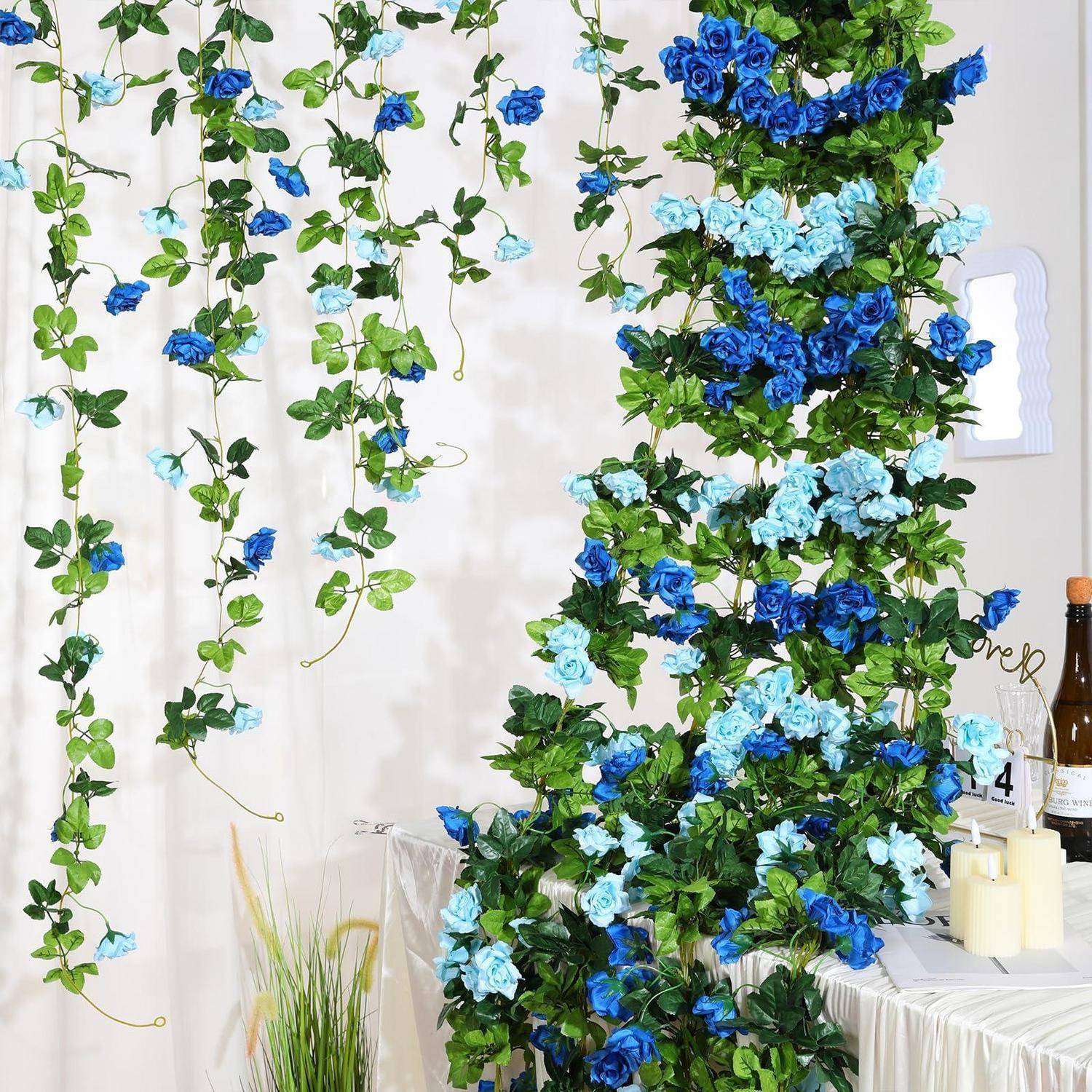 Blaue und hellblaue Blumengirlanden hängen an einer weißen Wand herab, neben einer hohen Blumensäule, mit Kerzen auf einem kleinen Tisch darunter.