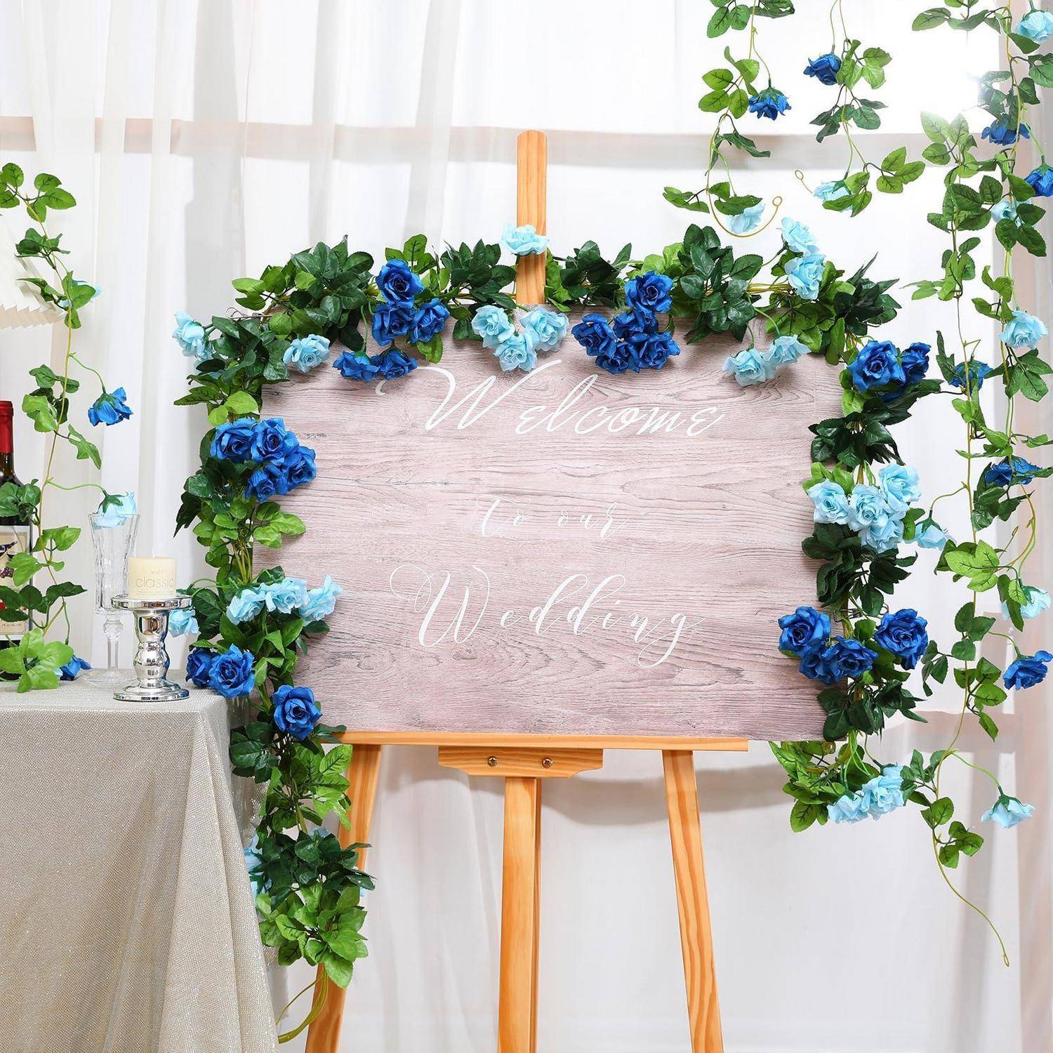 Willkommensschild zur Hochzeit auf einer Staffelei, umrahmt mit blauen und weißen Blüten und Grünpflanzen, mit der Aufschrift "Willkommen"" und ""Zu unserer Hochzeit"".