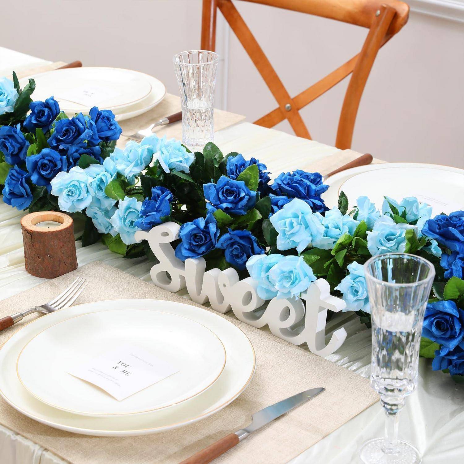 Elegante Tischdeckung mit blumigem Mittelstück in Blau und weißem „Sweet"-Schild, eingedeckt mit Tellern, Gläsern, Besteck und Servietten auf einem Esstisch.