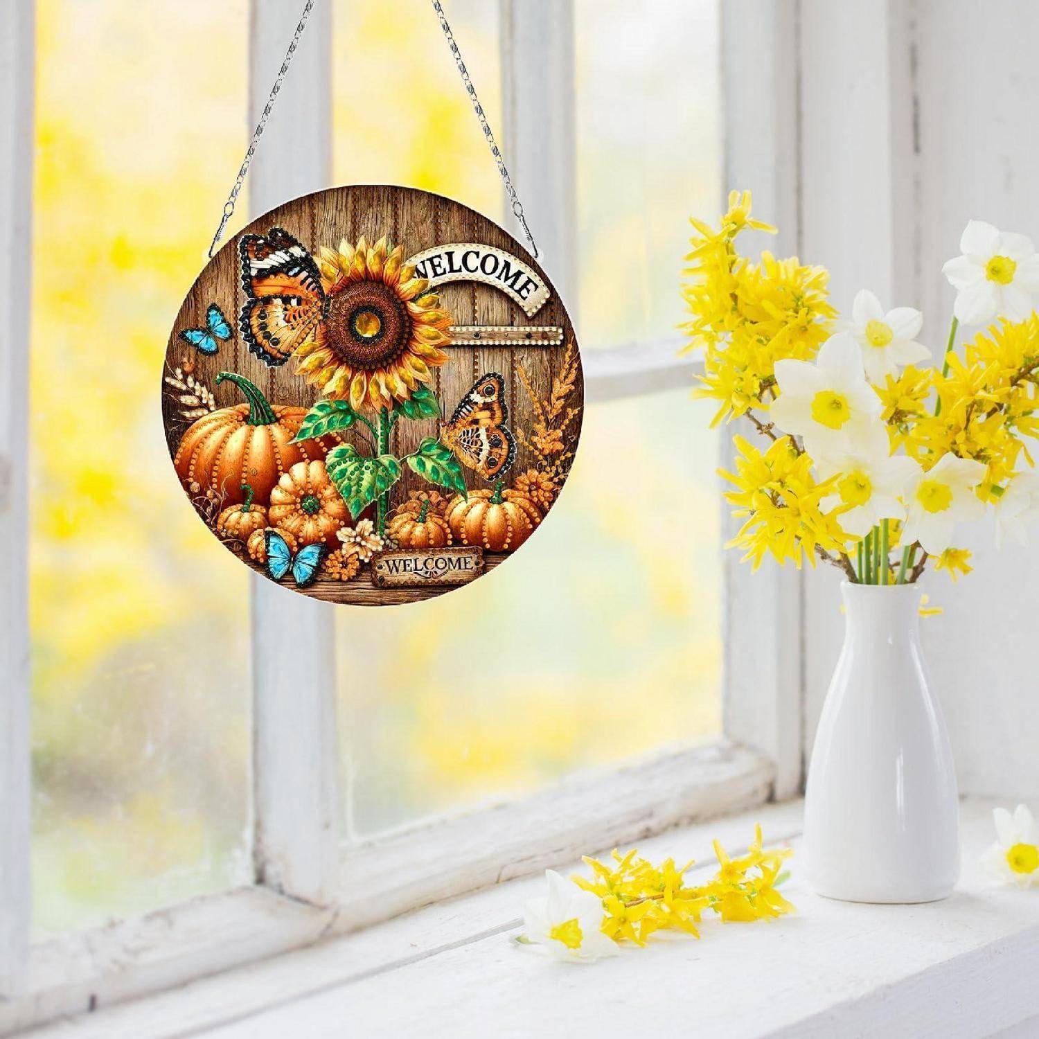 Ein rundes Herbst-Willkommensschild mit Kürbissen, Sonnenblume und Schmetterlingen hängt in einem hellen Fenster neben einer weißen Vase mit gelben und weißen Blumen.