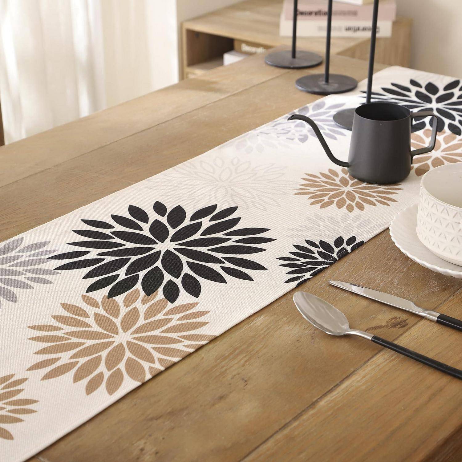 Esstisch mit einem Tischläufer mit Blumenmuster, schwarzem Krug, weißer Tasse und Besteck auf einer Holzoberfläche.