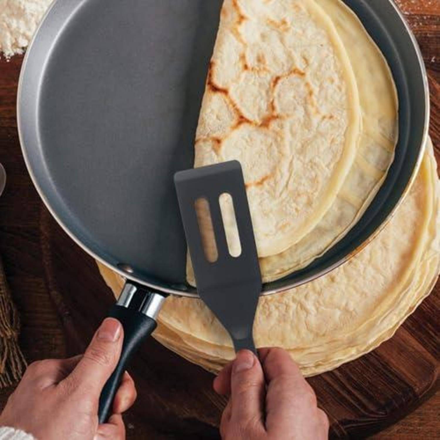 Hände verwenden einen Spatel, um einen Crepe aus einer Antihaftpfanne zu heben, mit einem Stapel gekochter Crepes auf einer Holzoberfläche.