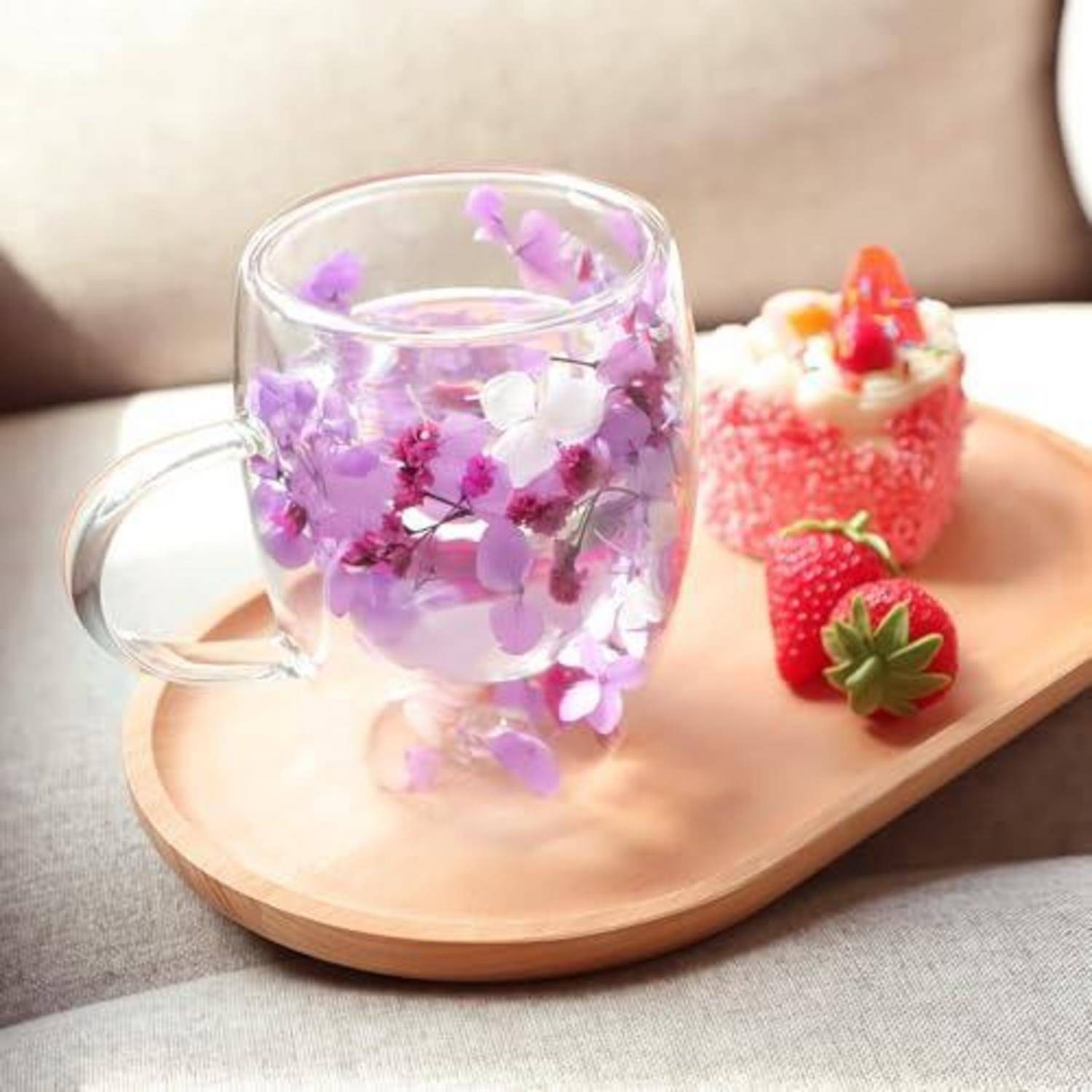 Glaskrug mit violetten Blumen auf einem Holztablett neben einem Dessert mit Erdbeeren und frischen Erdbeeren auf einem Sofa.