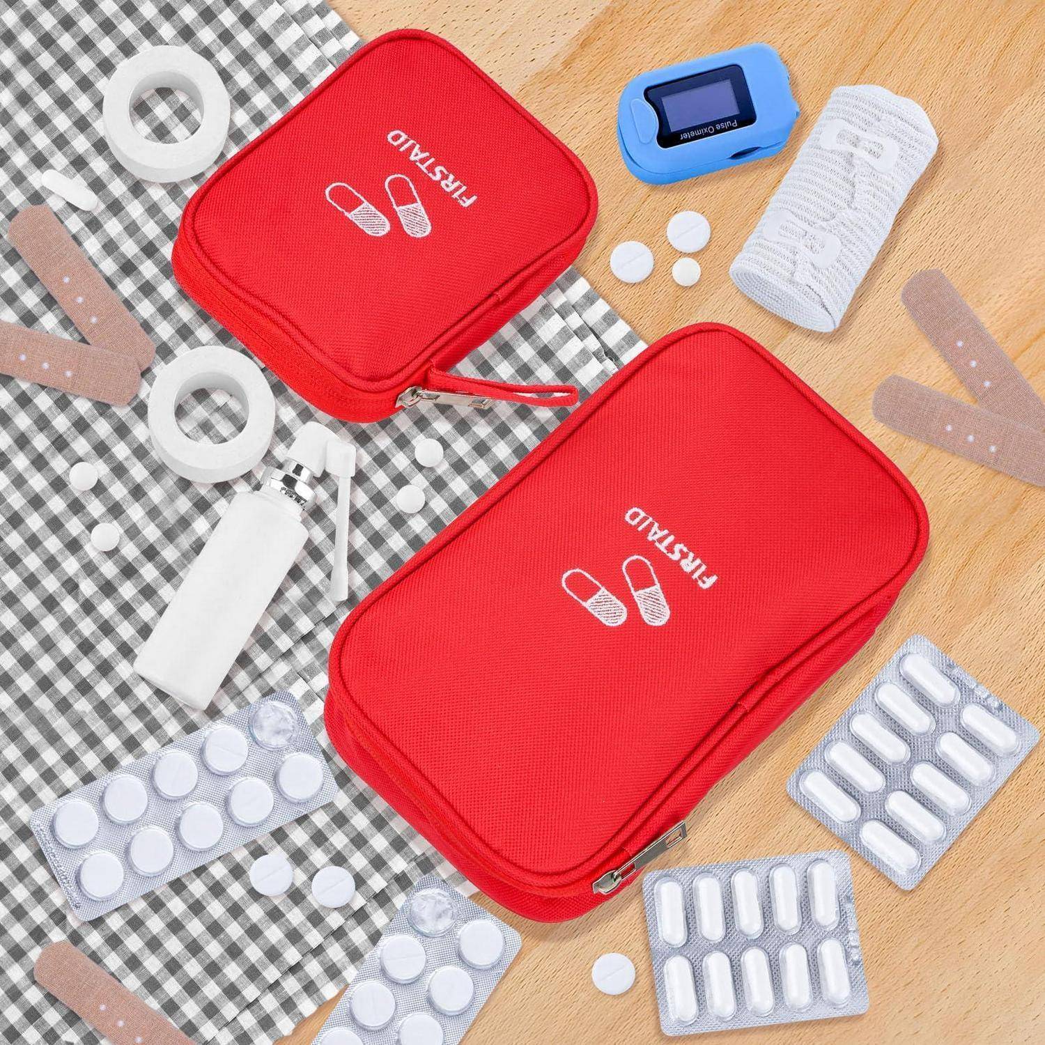 Zwei rote Erste-Hilfe-Kulturbeutel auf einer karierten Matte, umgeben von Verbandsmaterial, Tabletten, Gaze, Klebeband und einem blauen Pillendose-Organizer.