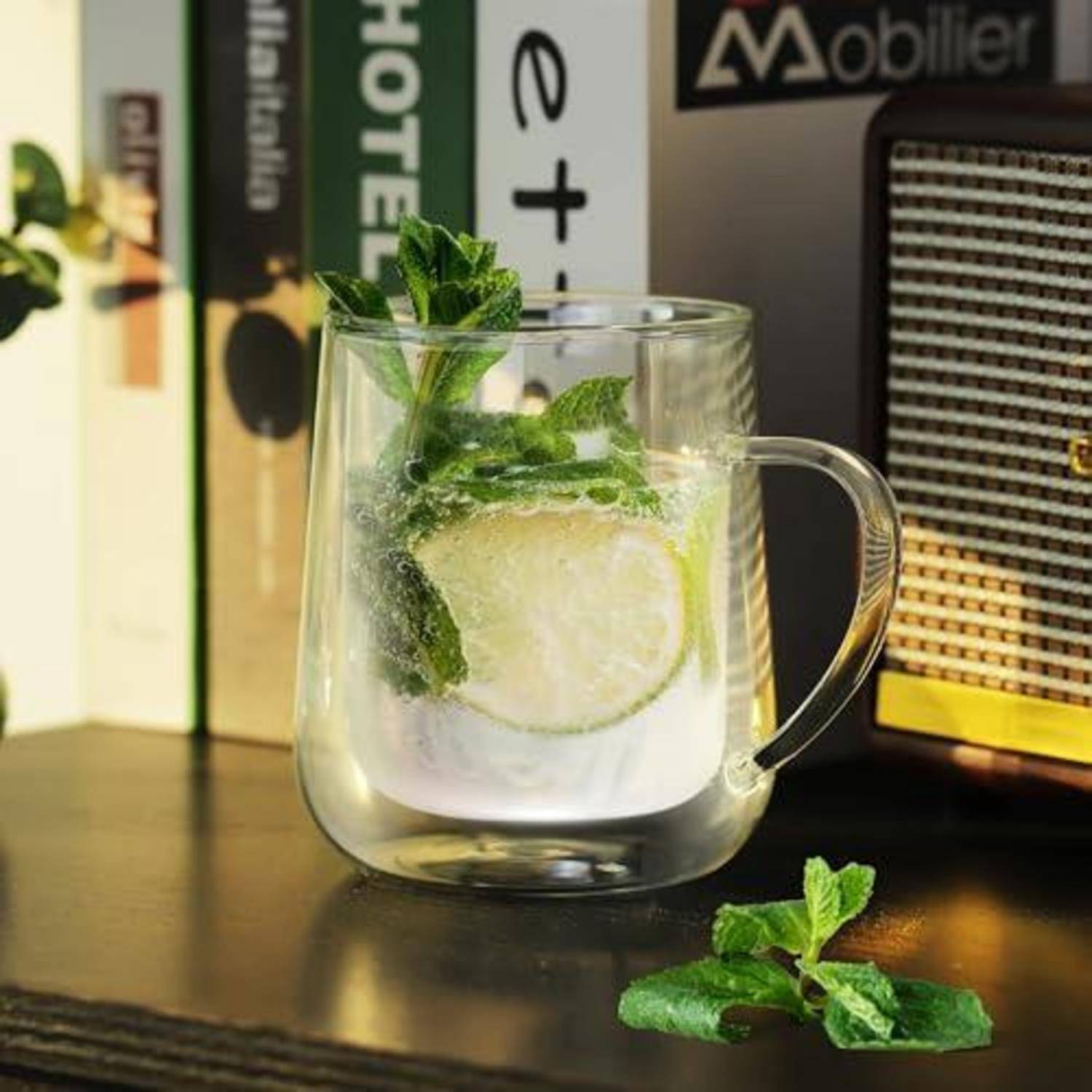 Glasbecher mit eiskaltem Zitronen-Minz-Getränk auf einem dunklen Tisch, mit einer Limettenscheibe und Minzspröslingen, neben Büchern und einem Radio angeordnet.