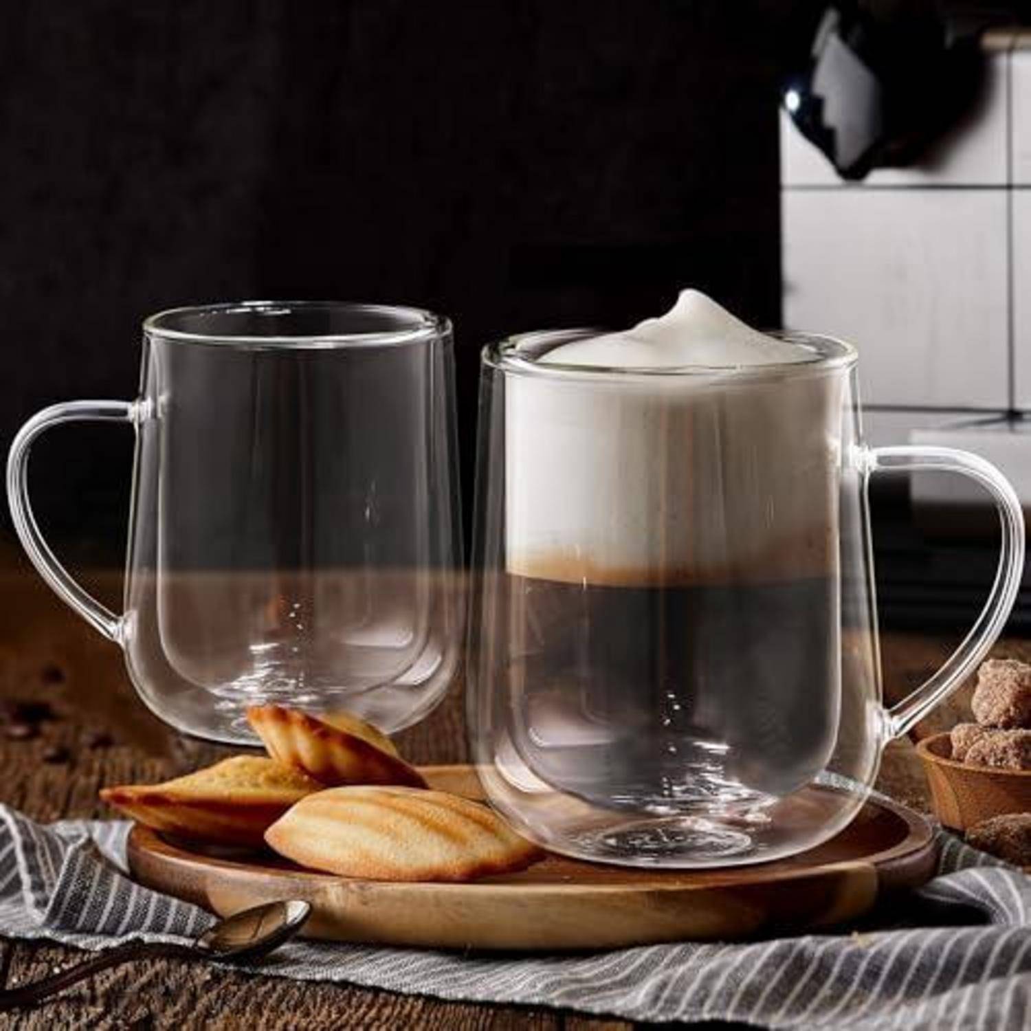 Zwei klare Glastassen auf einem Tablett, eine gefüllt mit geschichtetem Kaffee und Schaum obenauf und eine leere, daneben Kekse und ein Löffel auf einem rustikalen Tisch.
