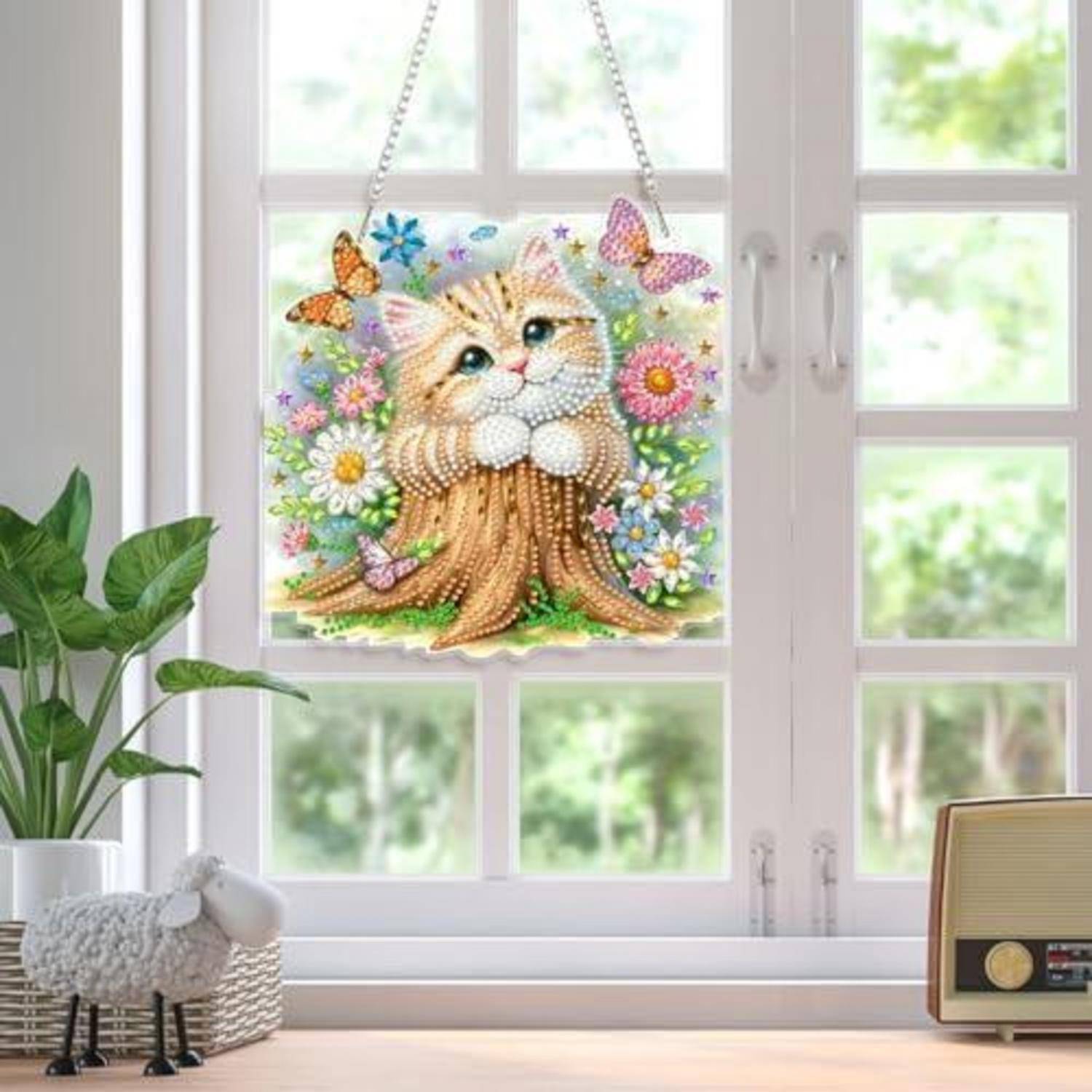 Diamantmalerei einer Katze, die aus einem Baumstumpf schaut, umgeben von Blumen und Schmetterlingen, hängend in einem sonnendurchfluteten Fenster.