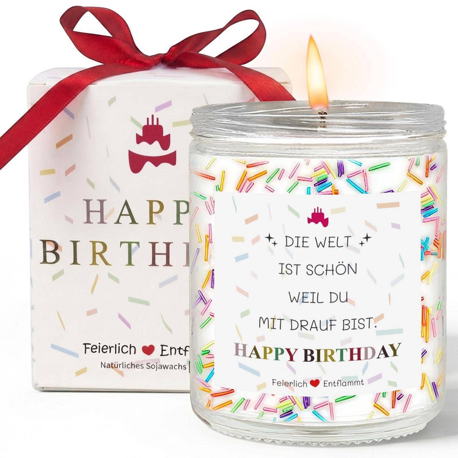 Brennende Geburtstagskerze in einem dekorierten Glas, mit einer Geschenkbox dahinter; das Etikett lautet „Die Welt ist schön weil du mit drauf bist. Happy Birthday