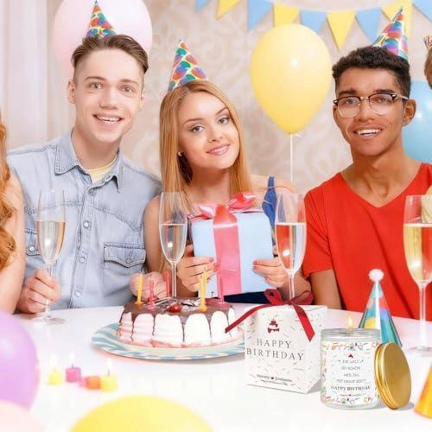 Drei Freunde mit Partyhüten lächeln an einem Geburtstagstisch mit Kuchen, Geschenk, Sektgläsern und Luftballons.