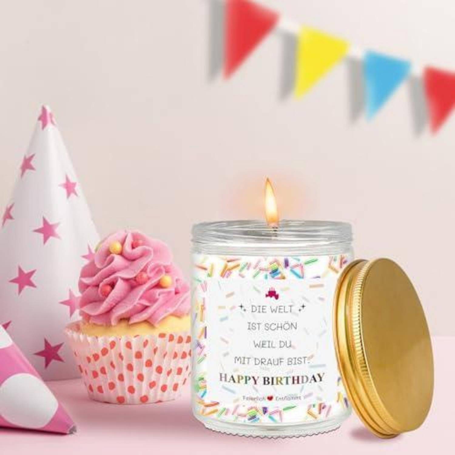 Eine Geburtstagsparty-Szene mit einer brennenden Kerze in einem dekorierten Glas mit der Aufschrift „ALLES GUTE ZUM GEBURTSTAG", einem rosa glasierten Cupcake und Partyhüten/Flaggen auf einem pastellfarbenen Hintergrund.