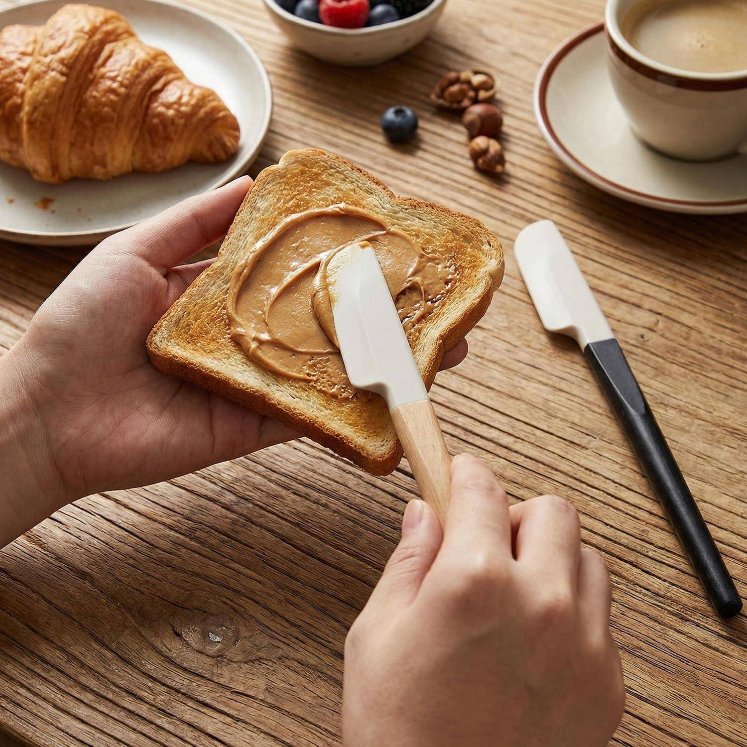 Hände verteilen Erdnussbutter auf Toast an einem Holztisch, mit einem Croissant, Kaffee und Nüssen im Hintergrund.