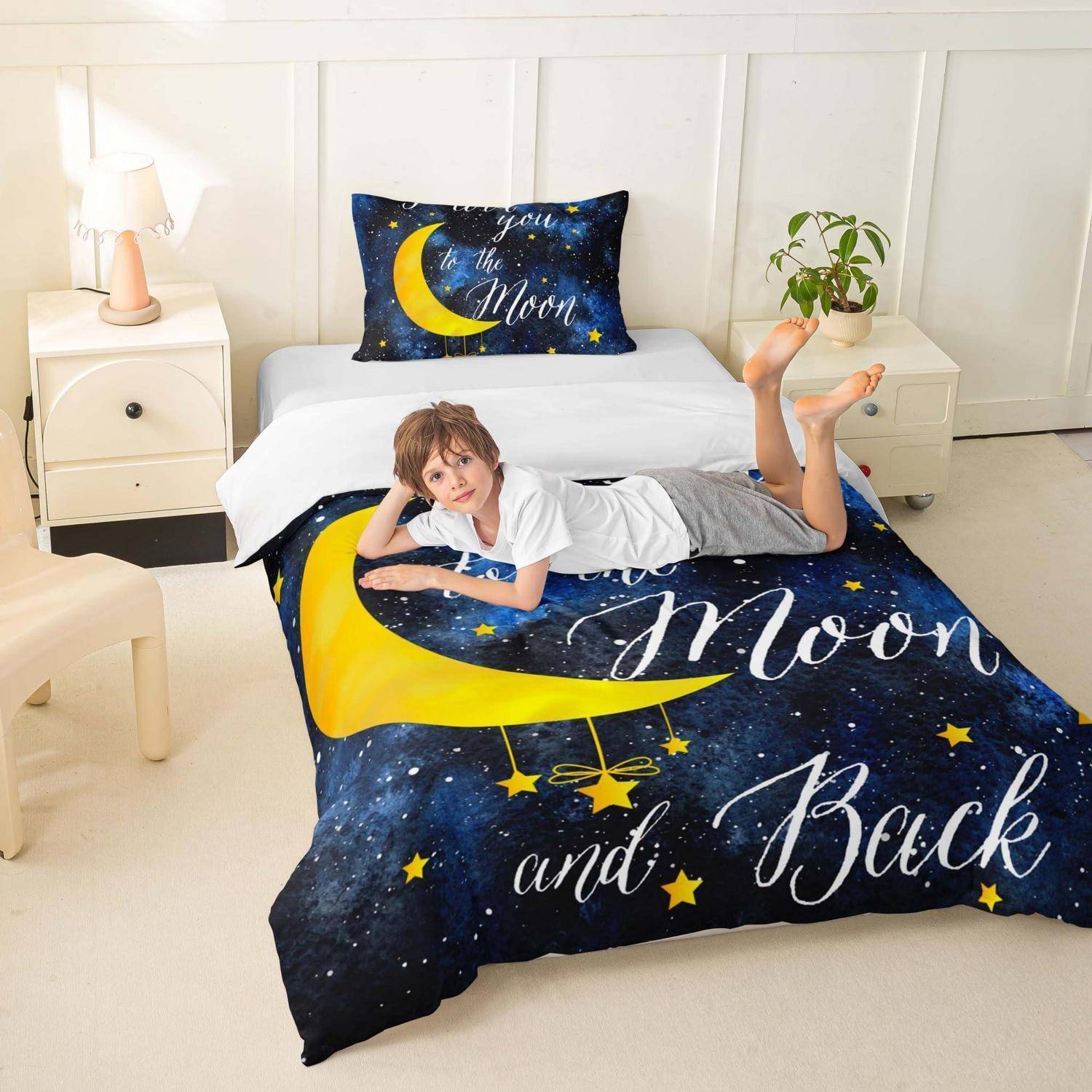 Kind, das auf einem Bett mit dunkelblauer Bettwäsche mit Mond- und Sternenmotiven liegt, auf der „To the Moon and Back" steht, in einem hellen Schlafzimmer mit Nachttischen und einer Lampe.
