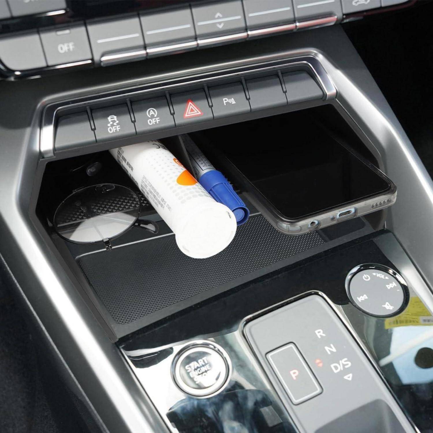 Ablageablage in der Mittelkonsole des Autos mit einem Smartphone, einer Röhre und einem Stift neben Tasten für Warnblinklicht und Parkassistent, über dem Schalthebel.