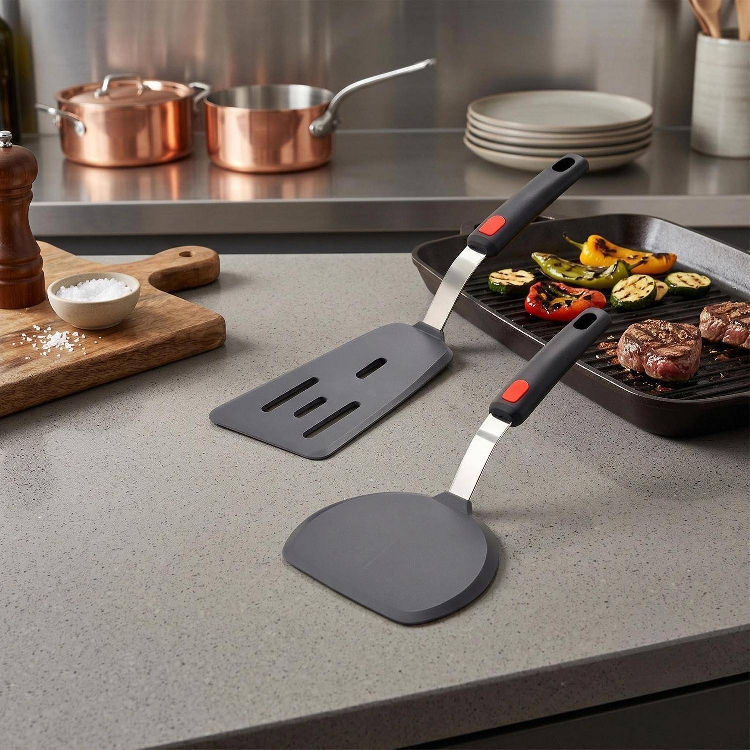 Zwei schwarze und silberne Spatel auf einer Küchenarbeitsplatte neben einer Grillpfanne mit Steaks und Gemüse, in einer modernen Kochszene.