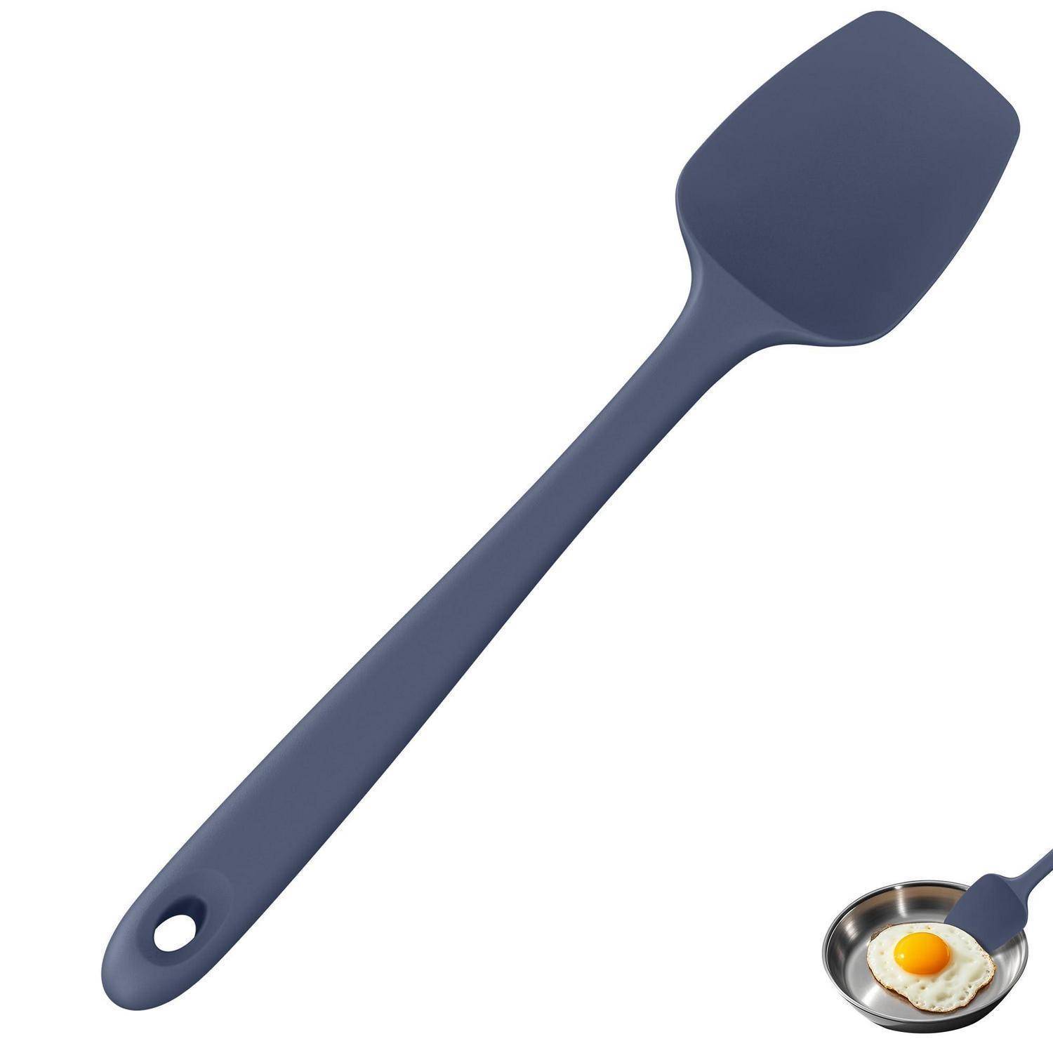 Dunkelblauer Silikonspatel mit Aufhängeloch, neben einer Bratpfanne mit einem Spiegelei gezeigt, was die Verwendung beim Kochen andeutet.